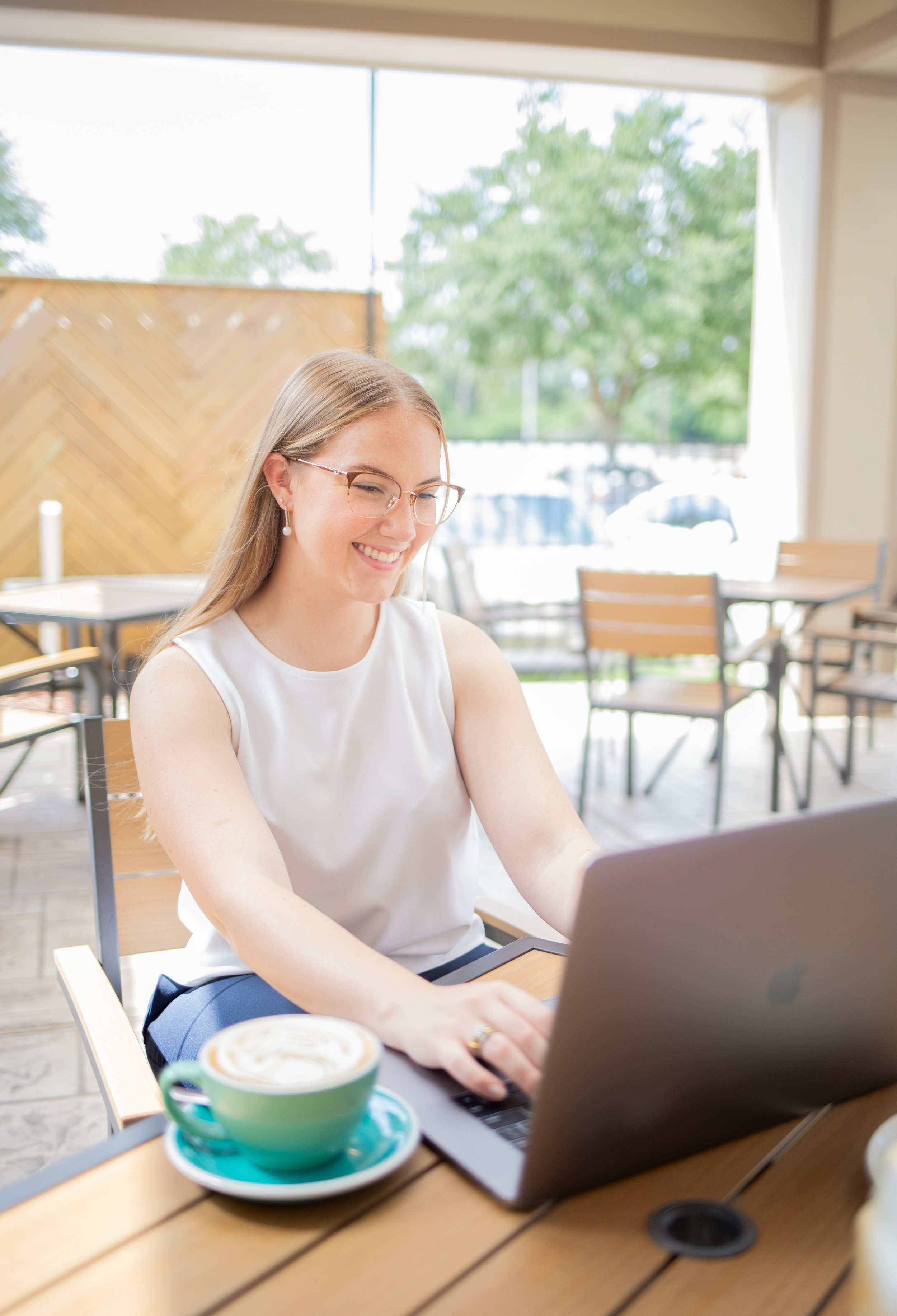 A young woman with glasses and pearl earrings is sitting at a table in a cafe, smiling while working on her laptop. A cup of coffee with latte art is in the foreground on a green saucer, and trees and parked cars are visible through large windows in the background.