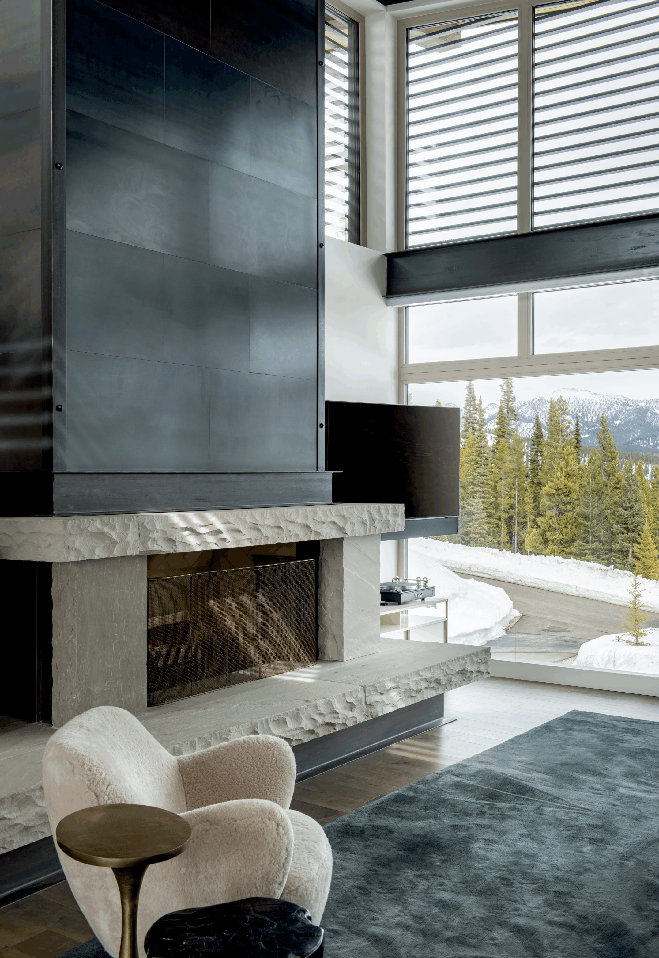 Modern living room with a stone fireplace, large windows showing a snowy mountain landscape, plush white chair, small side table, and a black round stool, with a dark gradient rug on wooden floor.