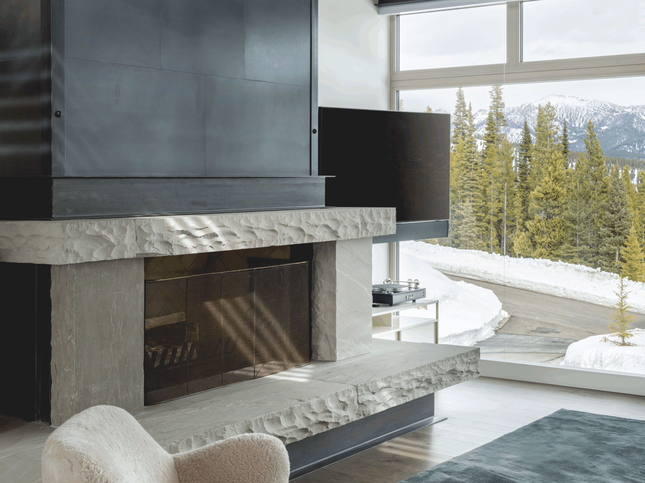 Modern mountain living room with a stone fireplace, large window showing snowy landscape and pine trees, and a cozy heather-colored armchair. Steel surround fireplace designed, fabricated, and built in Big Sky, Montana.