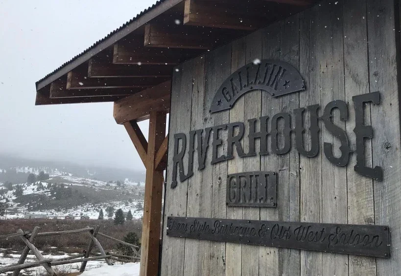 Wooden building with a custom steel and reclaimed lumber sign reading 'RIVERHOUSE Grill', in a snowy landscape.
