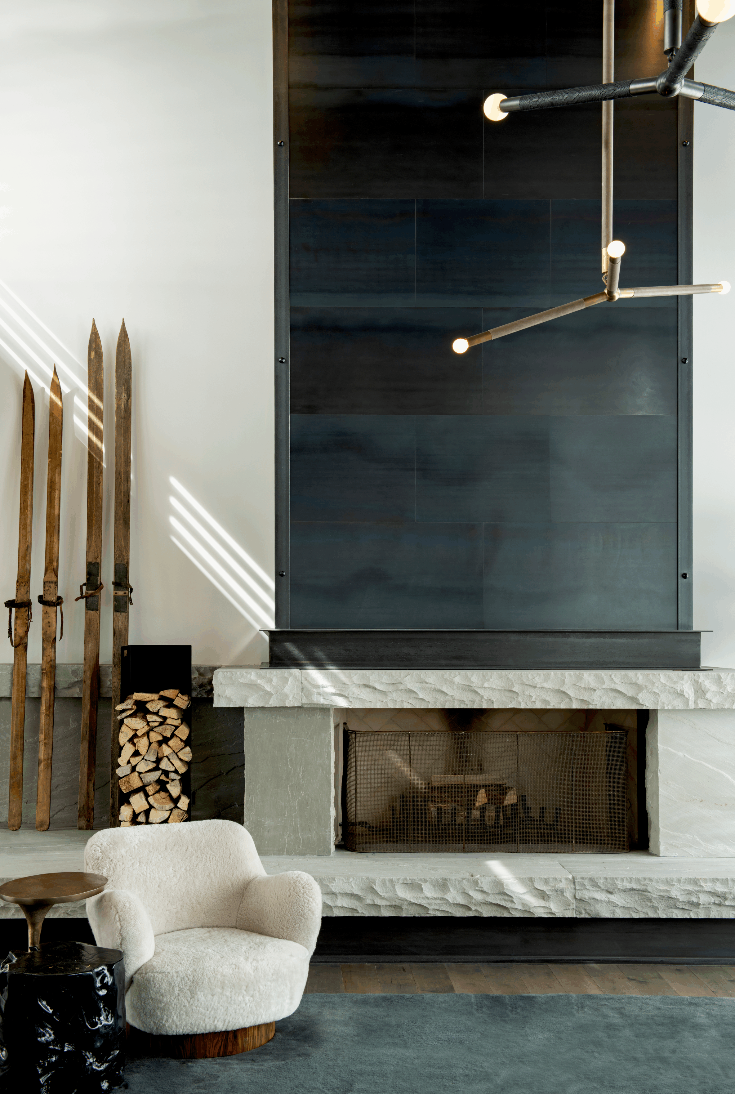 Living room with modern fireplace, stacked firewood, and wooden skis by the wall, with a plush white armchair and a small side table in the foreground.