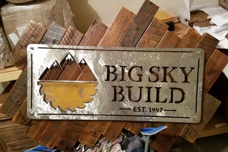 Metal sign that reads 'Big Sky Build, Est. 1997' with a mountain and sun logo, placed on a wooden surface.
