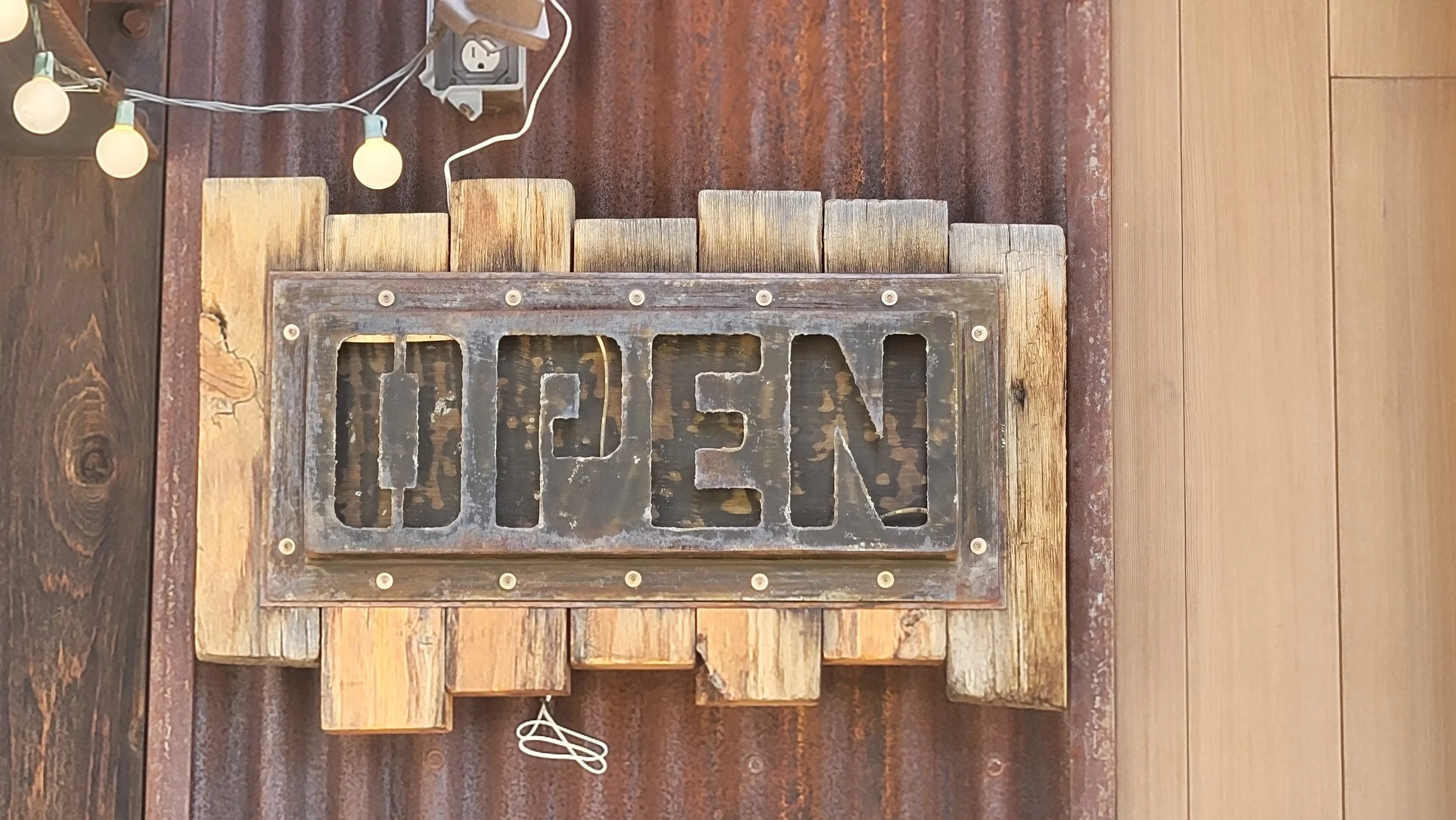 A rustic wooden sign with the word "OPEN" cut out, mounted on a steel wall.  "Open" sign is back lit and glows in the evening.