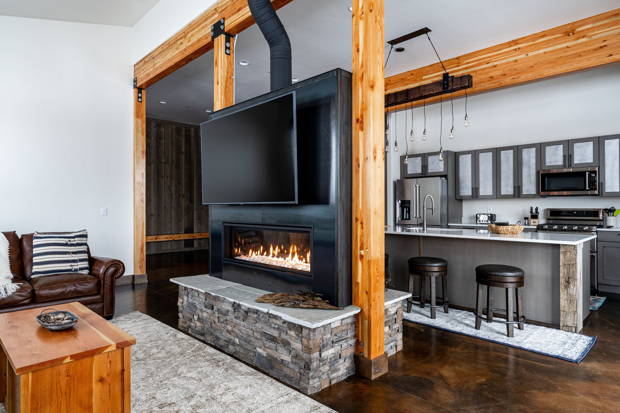 Living room with a leather sofa and a steel surround, 360 degree fireplace. Open kitchen with gray cabinets, stainless appliances, and an island with bar stools.  Steel fireplace surround and stonework completed by Jack Creek Contracting.