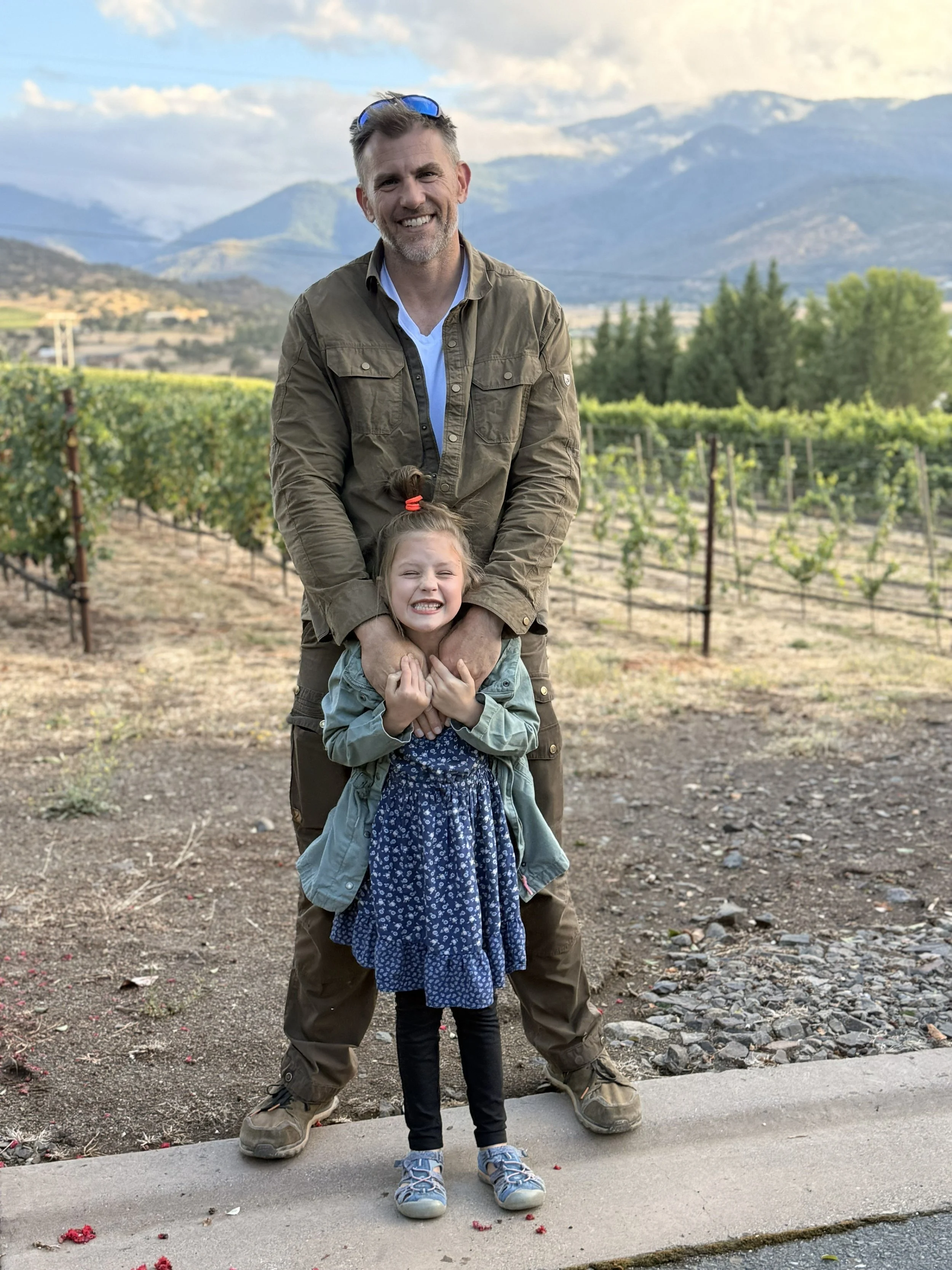 A man in outdoor clothing and sunglasses on his head stands behind a young girl, holding her shoulders. They are smiling broadly, with the girl showing her teeth and squinting. The background features vineyards, trees, mountains, and a partly cloudy sky.