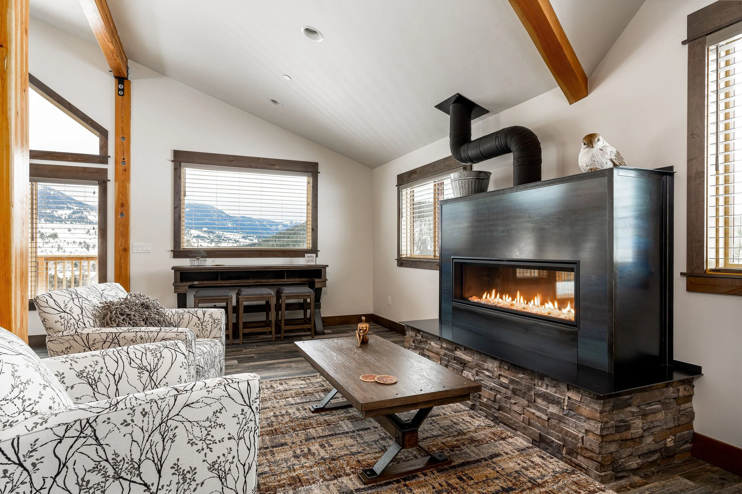 Living room with a rectangular wooden coffee table, a custom stone/steel fireplace with a black insert and a mounted bird sculpture, wooden beams, large windows showing mountain views. Fireplace surround and stonework by Jack Creek Contracting.