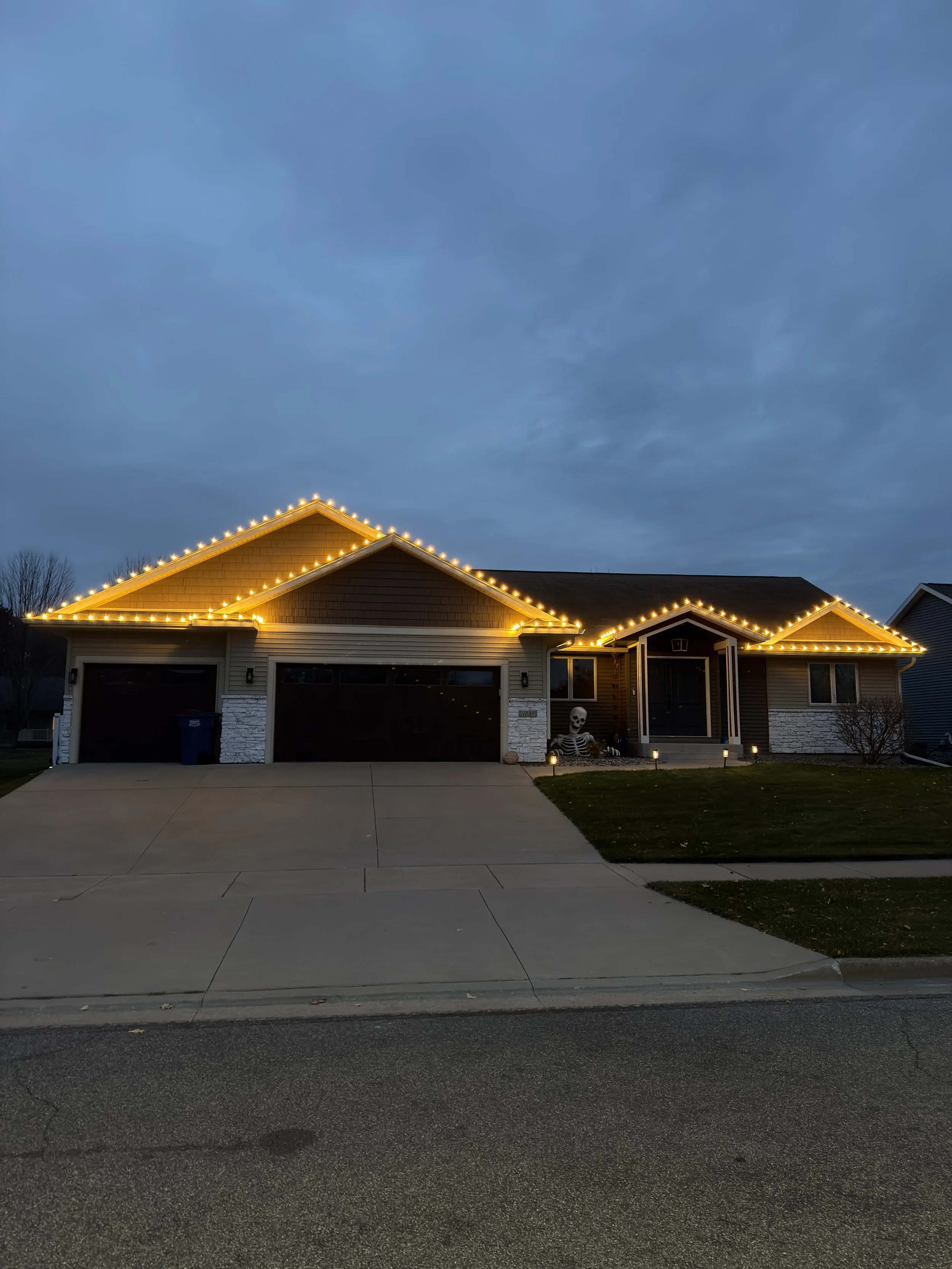A house with illuminated Christmas lights outlining the roofline during dusk or early evening, with a cloudy sky in the background and a skeleton Halloween decoration near the entrance.