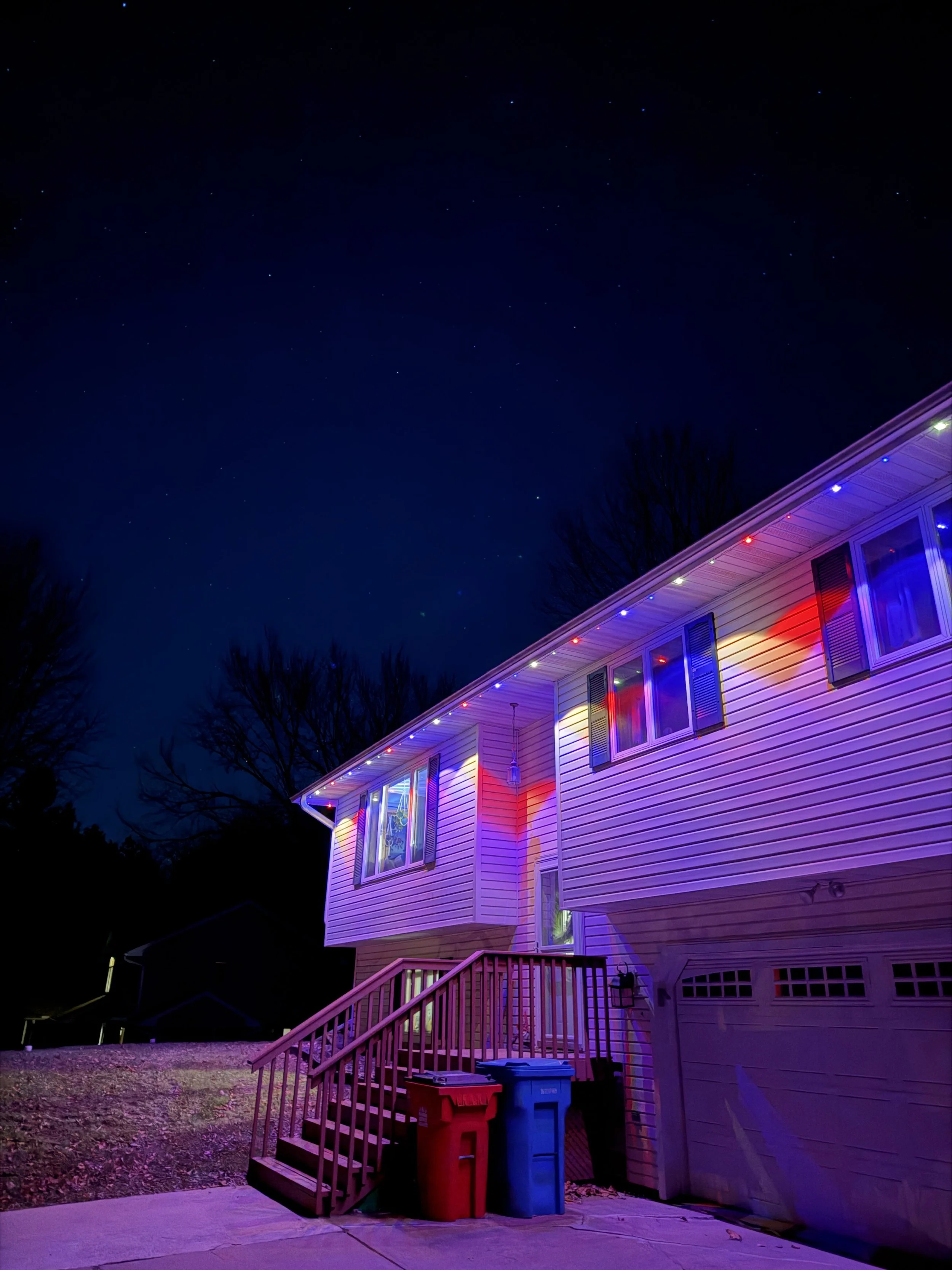 Nighttime view of a house decorated with colorful Christmas lights along the roofline. The house has siding and a front staircase leading to the entrance. There are trees in the background and trash bins near the stairs.
