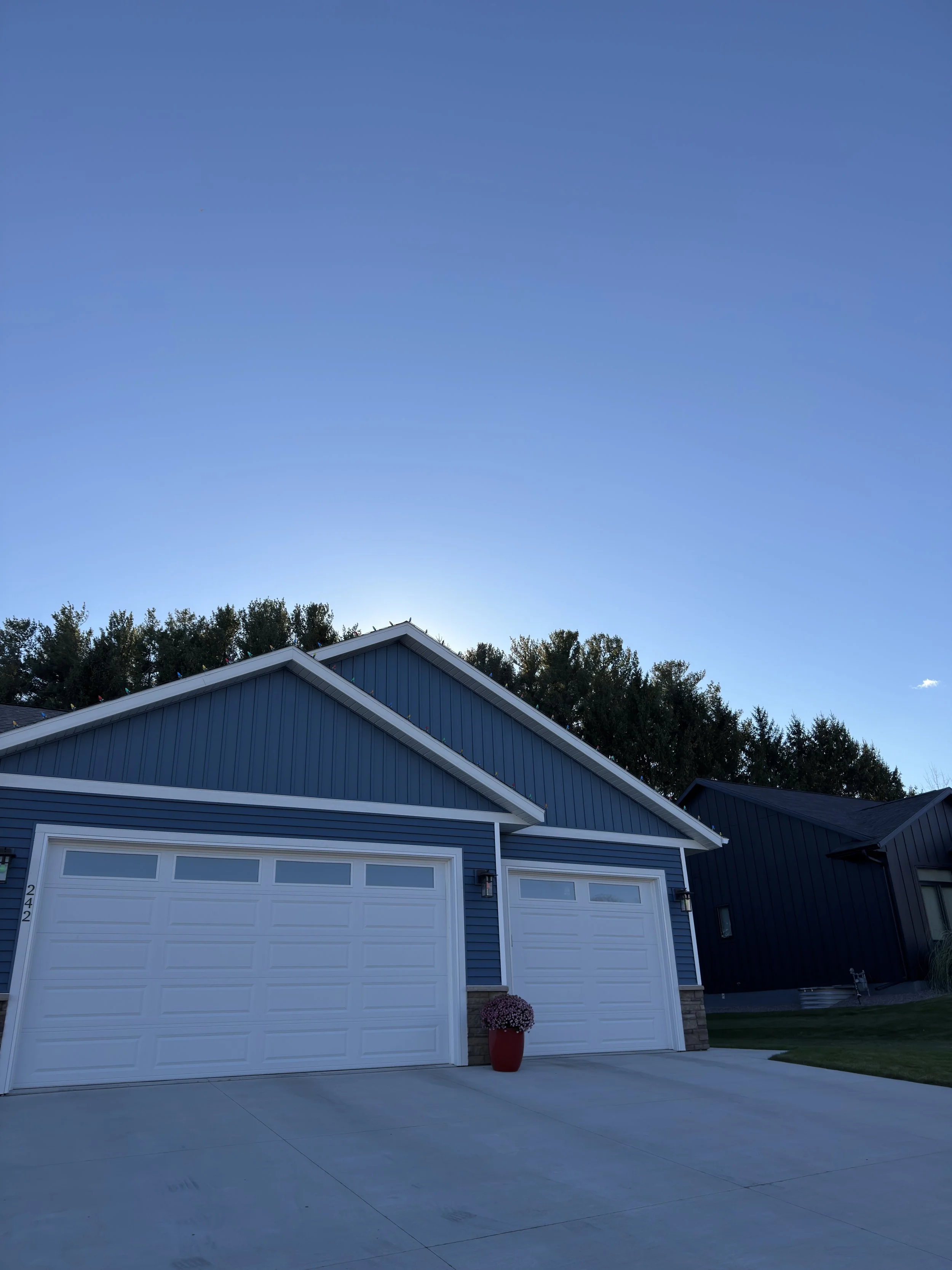 View of a blue house with a white garage door, a red flower pot with pink flowers, and a clear blue sky above.