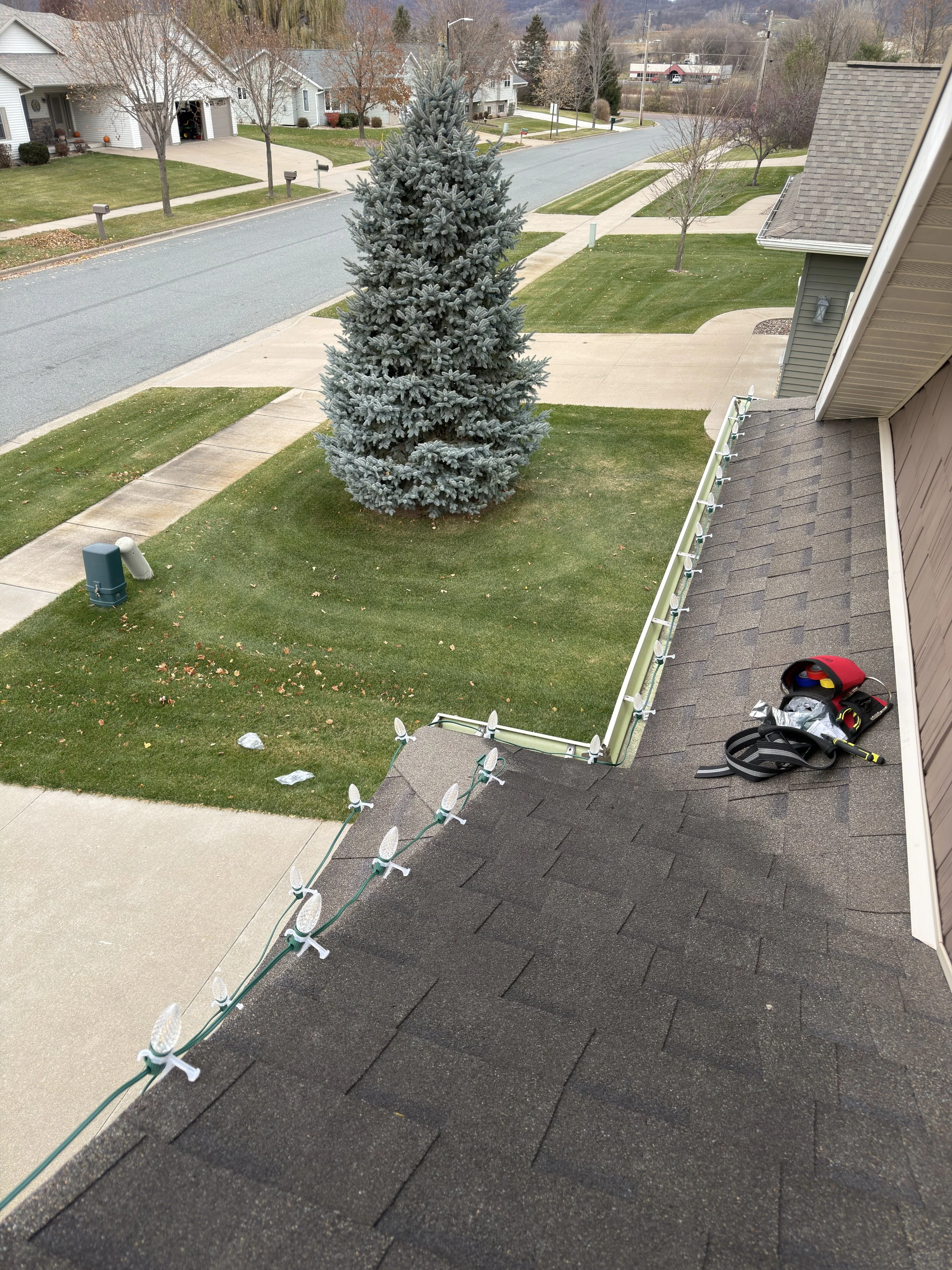 The photo shows a view from a house looking towards the front yard and street. There are Christmas lights installed on the edge of the roof, and a toolbox with tools on the patio. A large evergreen tree, several leafless trees, and houses on the street are visible, with a backdrop of distant hills or mountains.