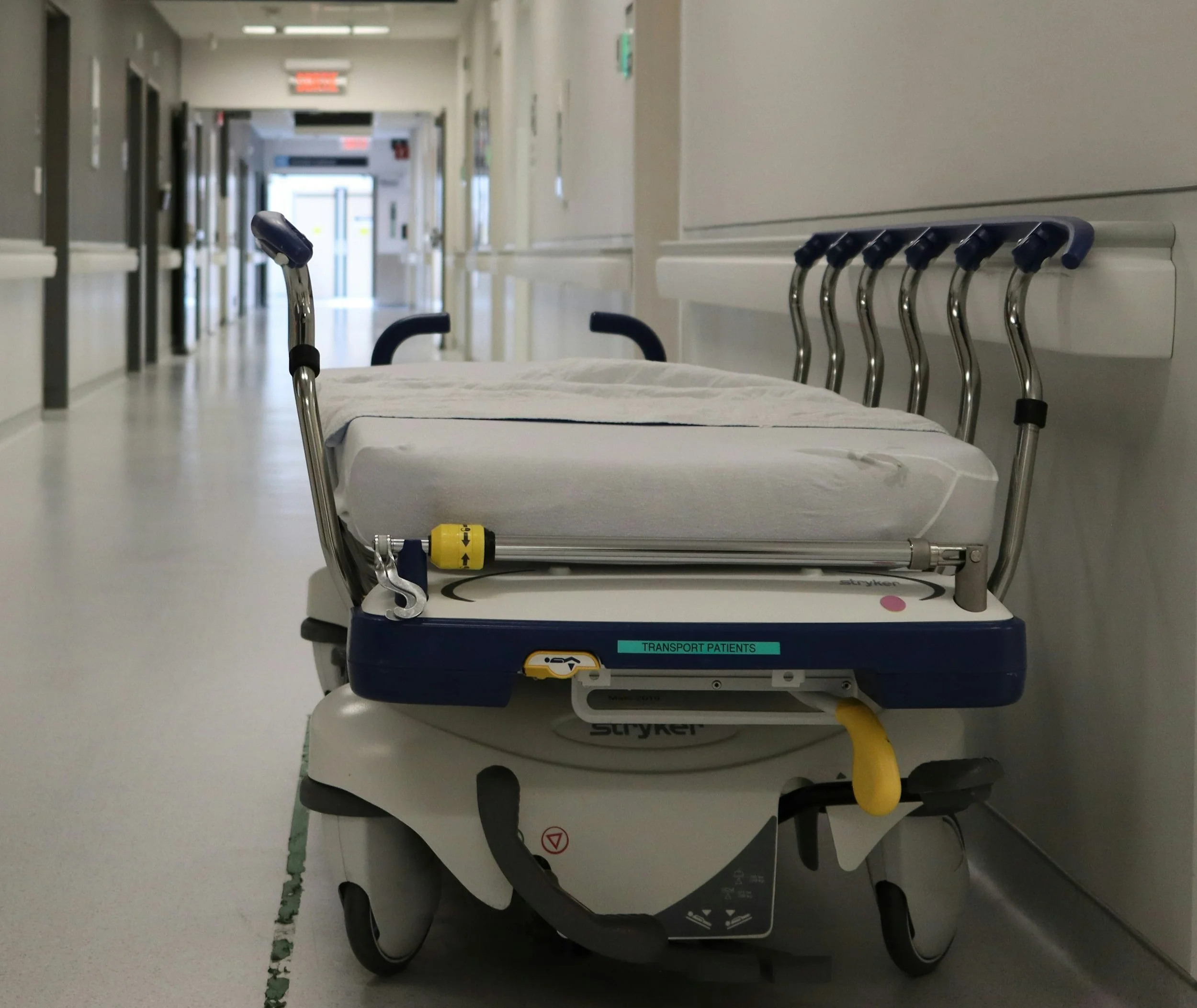An empty hospital stretcher with black handles and a yellow safety lock, positioned in a corridor with medical equipment and doors.