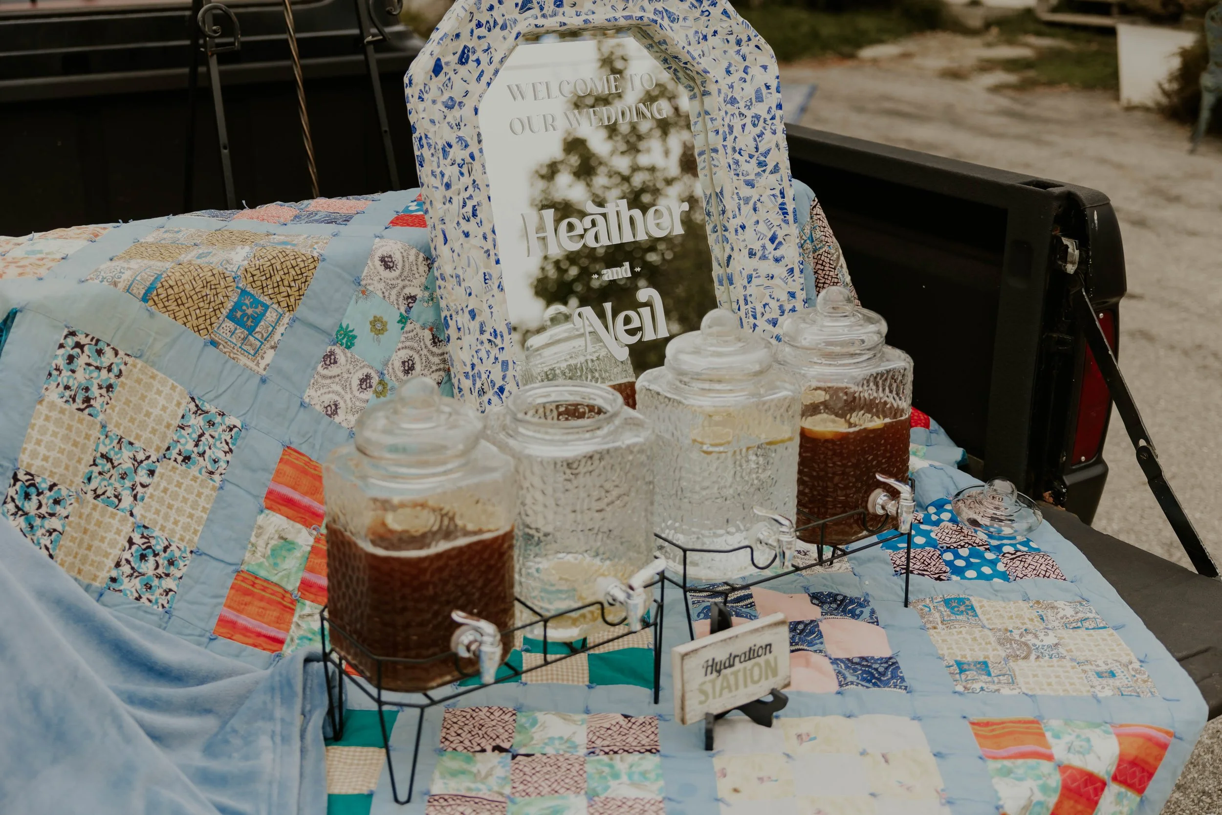 A wedding hydration station setup on a table covered with a patchwork quilt. The table displays glass jars filled with different drinks, and a mirror sign reads, 'Welcome to our wedding Heather and Neil.' The background shows a trailer and outdoor su