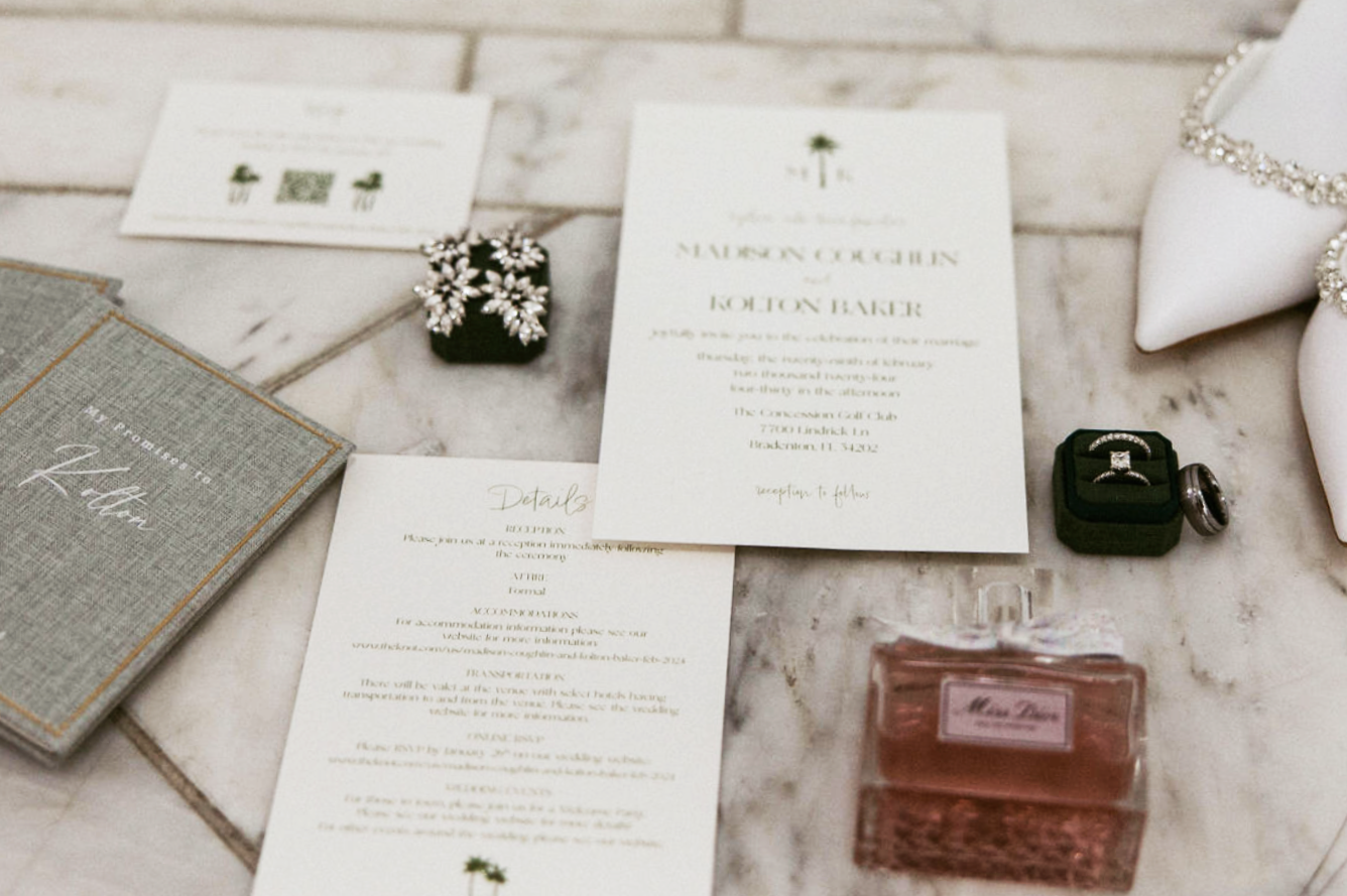 Wedding invitation and rings on a marble table, with perfume and jewelry nearby.
