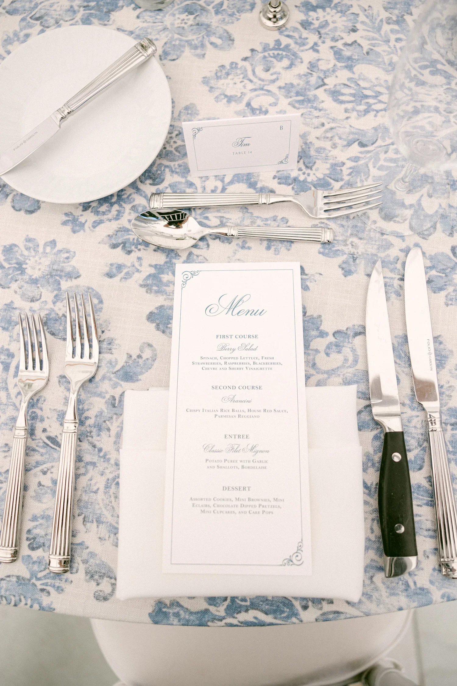 Table setting with cutlery, a menu, a table card labeled 'Tim', and a glass of water on a blue and white patterned tablecloth.