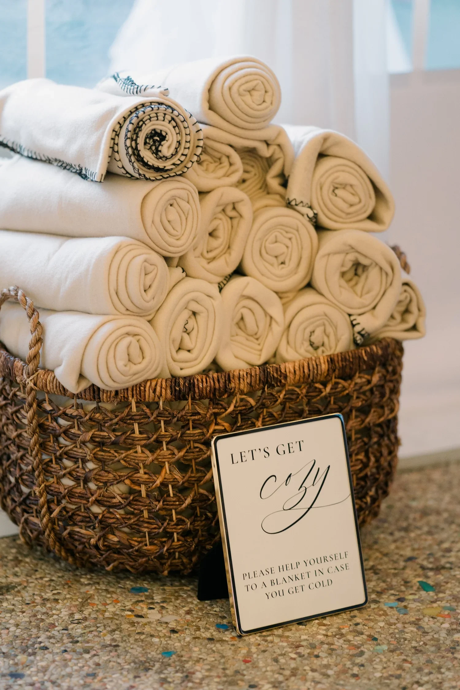 A woven basket filled with rolled white towels with decorative black stitching, placed on a speckled countertop. A sign in front reads, 'Let's get cozy. Please help yourself to a blanket in case you get cold.'