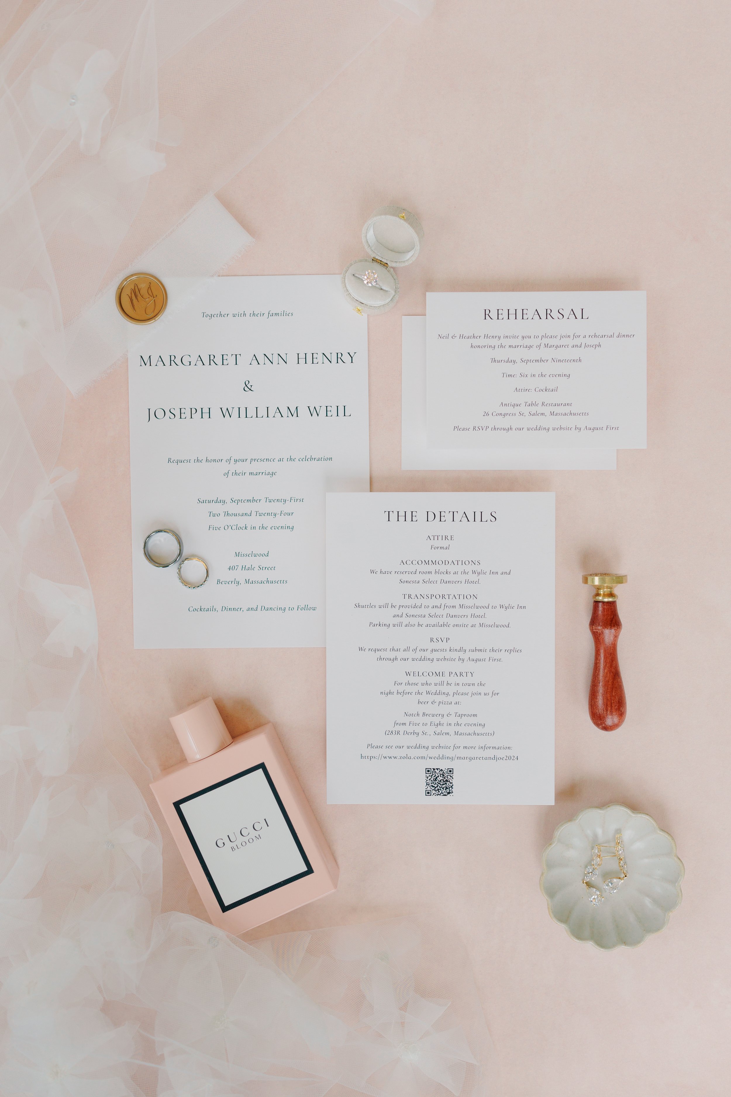 Wedding invitation cards, rings, and jewelry arranged on a pink surface with a Gucci Bloom perfume bottle and a small shell-shaped dish.