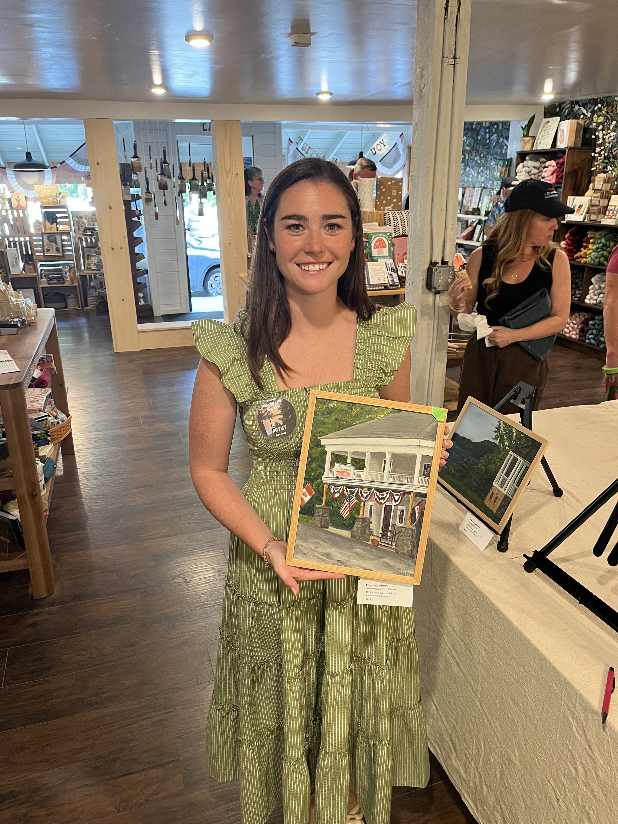 A young woman with long brown hair in a green dress holding a painting of a house with a porch, standing inside a shop or gallery with other artworks and people in the background.