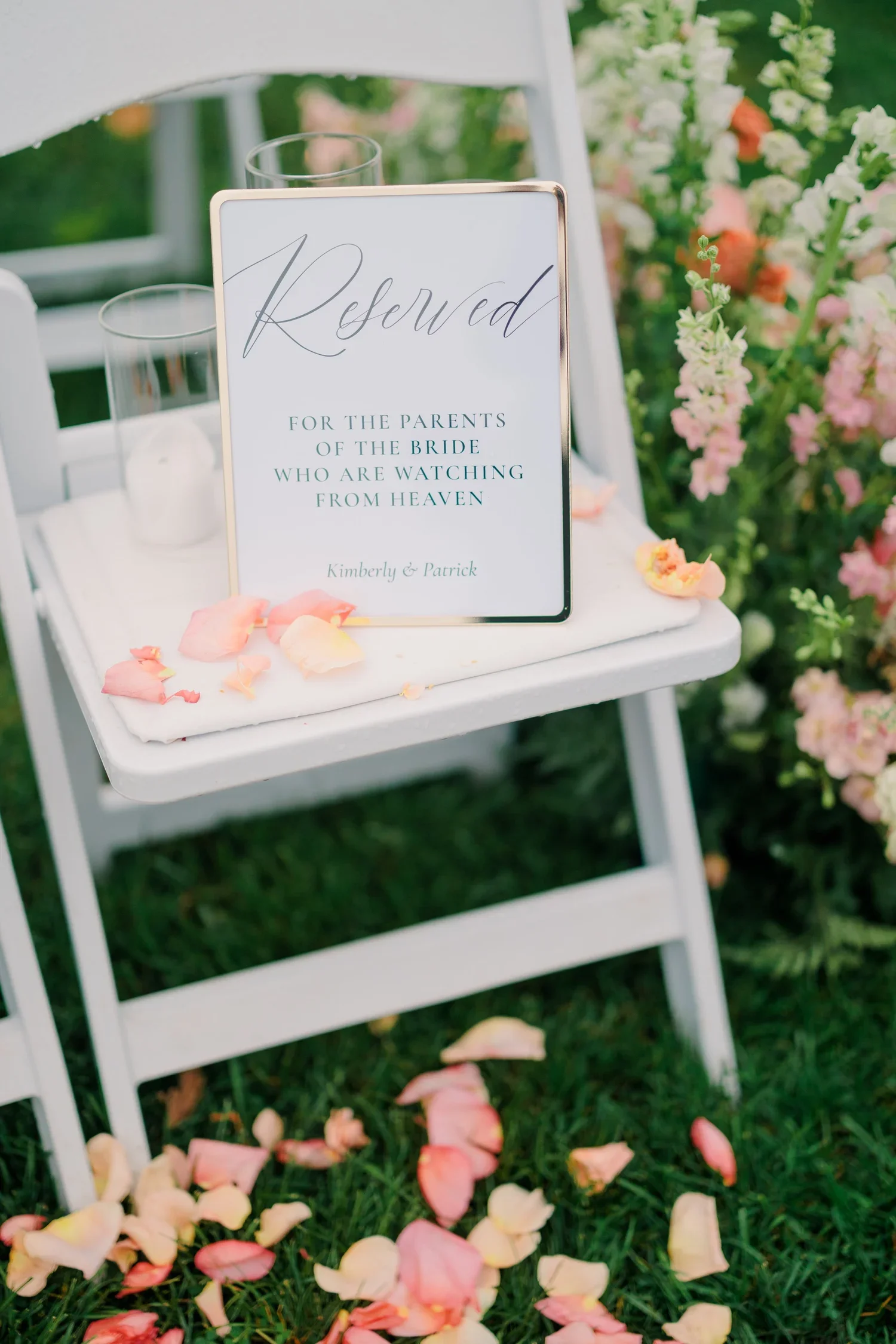 White wooden chair with a 'Reserved' sign, surrounded by pink and cream rose petals on grass, and colorful flowers in the background.