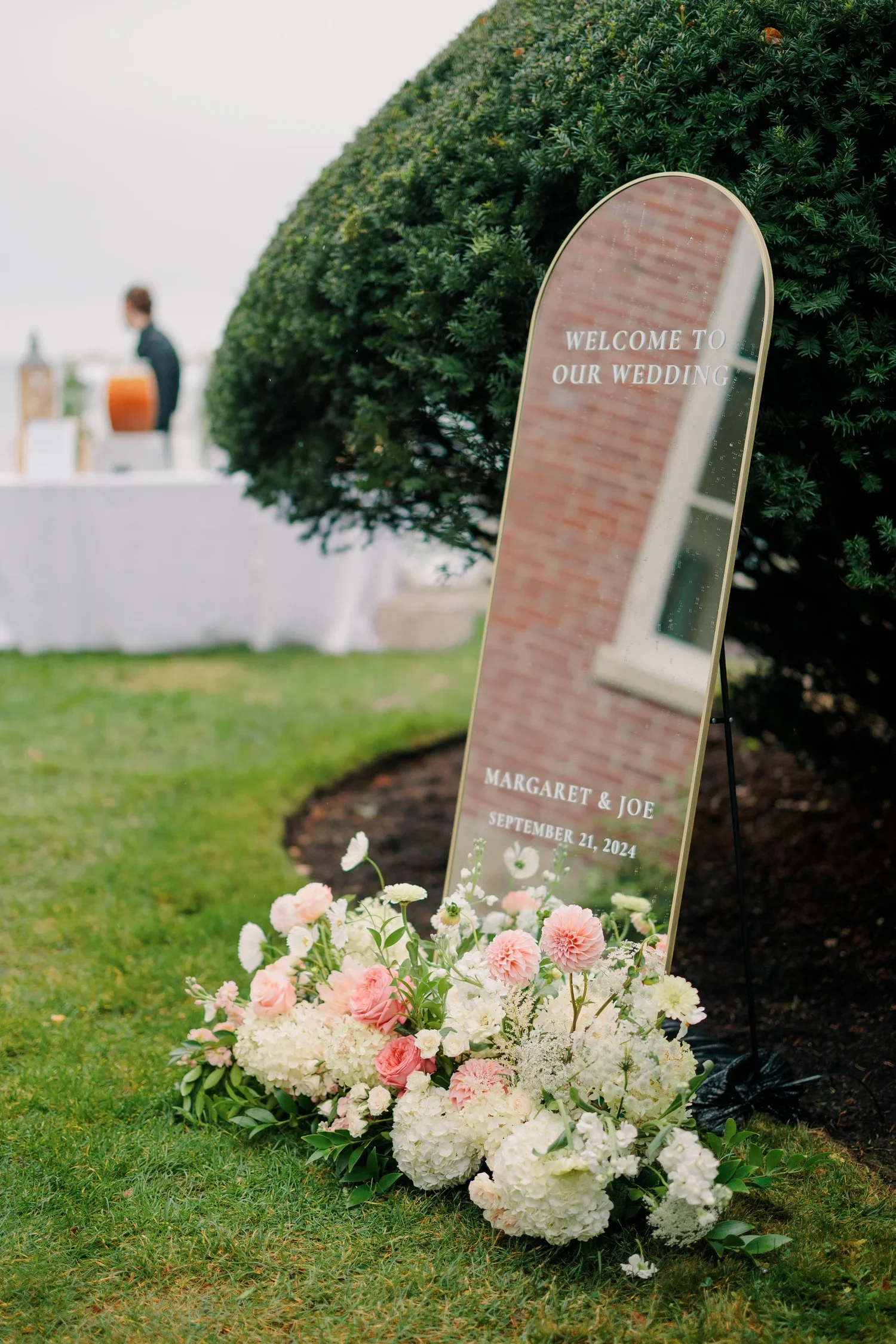 A wedding sign with the text "Welcome to our wedding," names Margaret and Joe, and the date September 21, 2024. The sign is placed outdoors next to a lush green bush, decorated with a bouquet of pink and white flowers at the base.
