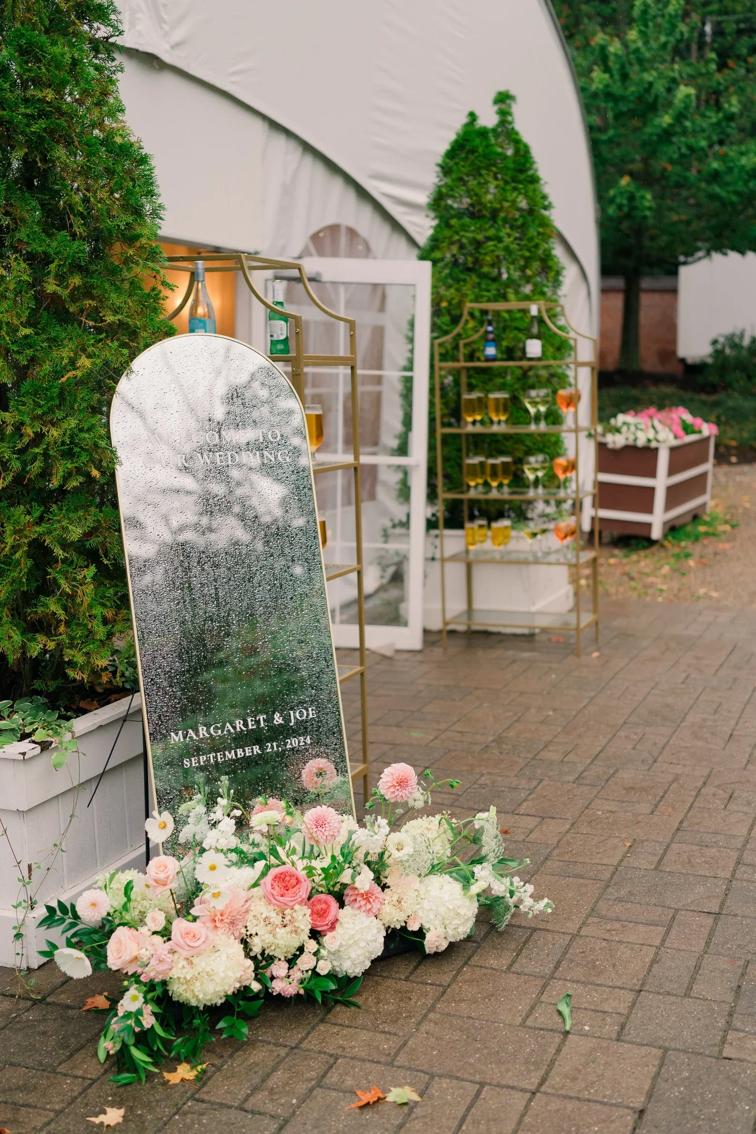 Wedding sign with floral arrangement and decorative bottles, reading "Welcome to Our Wedding, Margaret & Joe, September 21, 2024," located outdoors with trees and a floral display in the background.