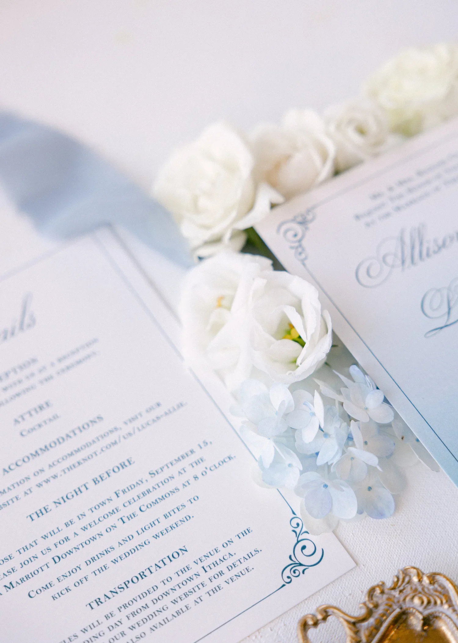 White wedding invitation card with elegant blue script and decorative blue design, surrounded by white roses and gold ornate decorations.