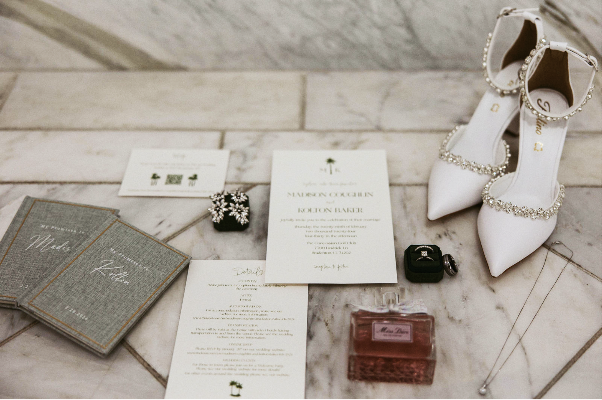A pair of white wedding shoes with rhinestone embellishments, wedding invitation and programs, a small box with jewelry, and a pink perfume bottle are arranged on a light-colored tiled floor.