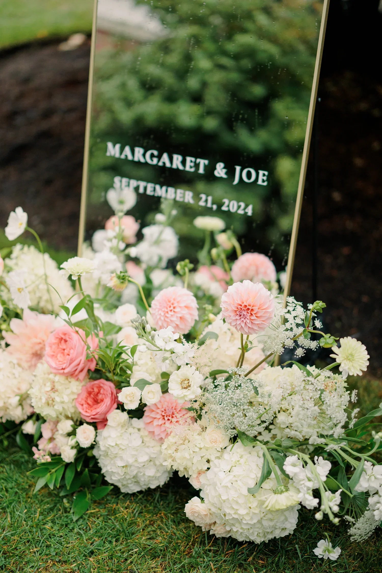 A floral arrangement at a memorial with a glass placard displaying the names Margret & Joe and the date September 21, 2024. The flowers include white hydrangeas, pink carnations, and various white and pink blooms surrounded by green foliage.