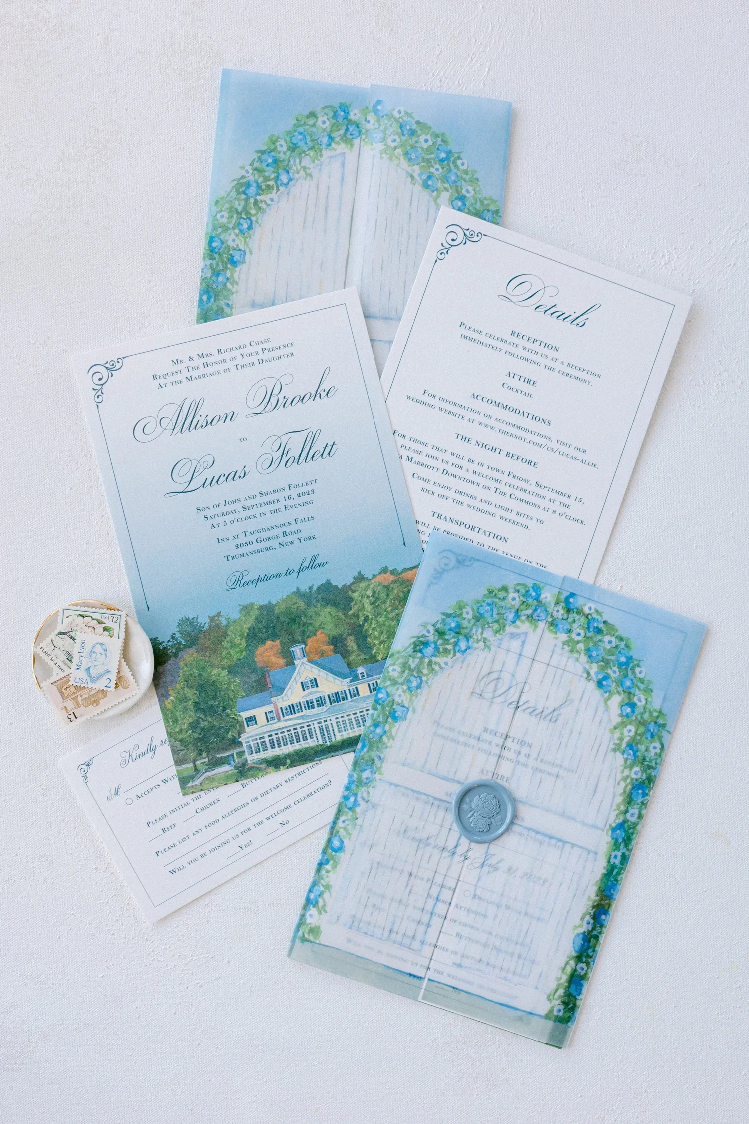 A collection of wedding invitations, RSVP cards, and a wax seal on a white surface. The invitations feature a blue floral design and a watercolor illustration of a house surrounded by trees.
