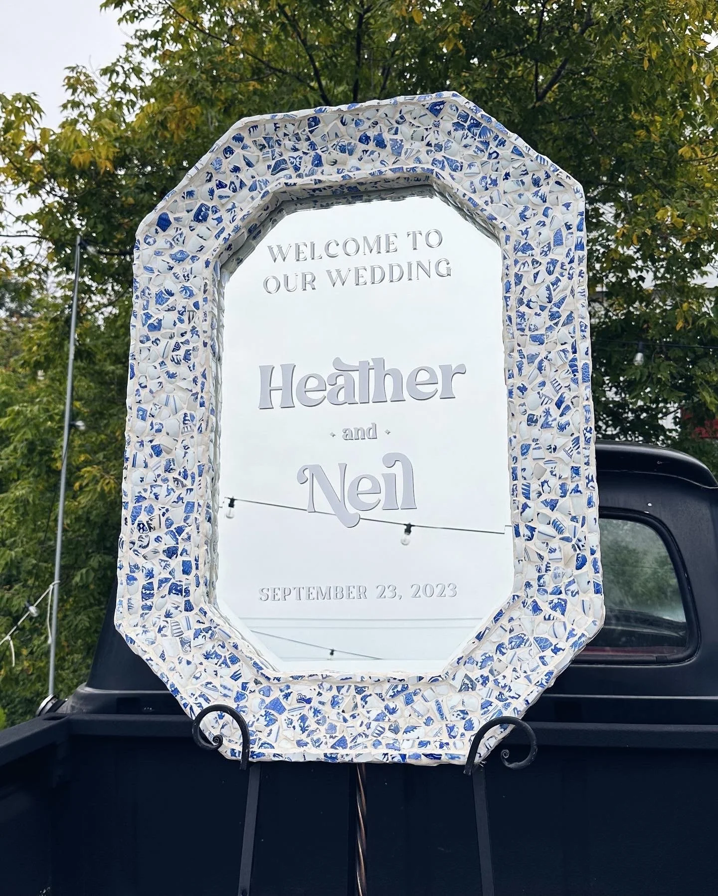 Wedding sign with a decorative white and blue border, reading 'Welcome to our wedding Heather and Neil September 23, 2023', mounted on a stand on a vehicle, with green trees in the background.