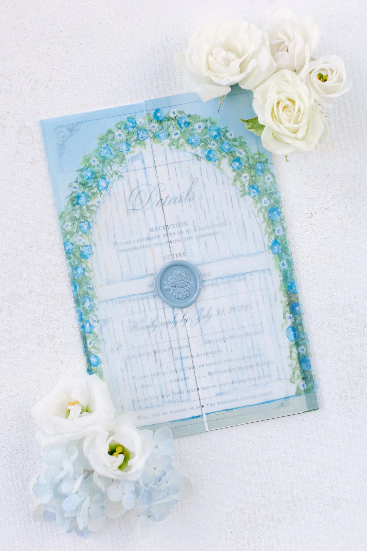 Wedding reception details card with a floral border, blue and white flowers, a blue wax seal, and white flowers on white fabric.