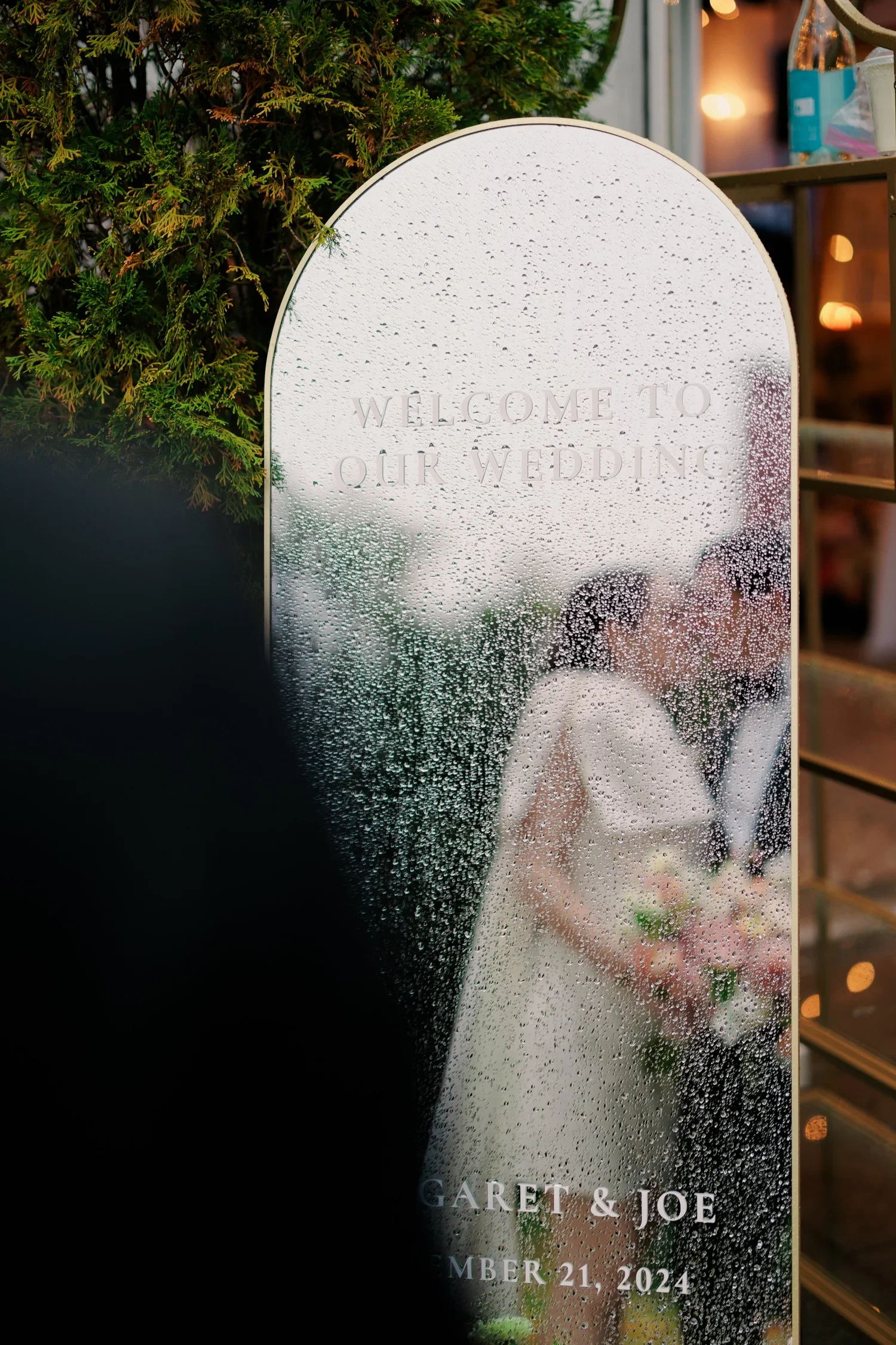 A foggy wedding reception sign with the message 'Welcome to Our Wedding' and the names Garrett & Joe, dated November 21, 2024, visible through a rain covered glass surface. Green shrubbery surrounds the sign.