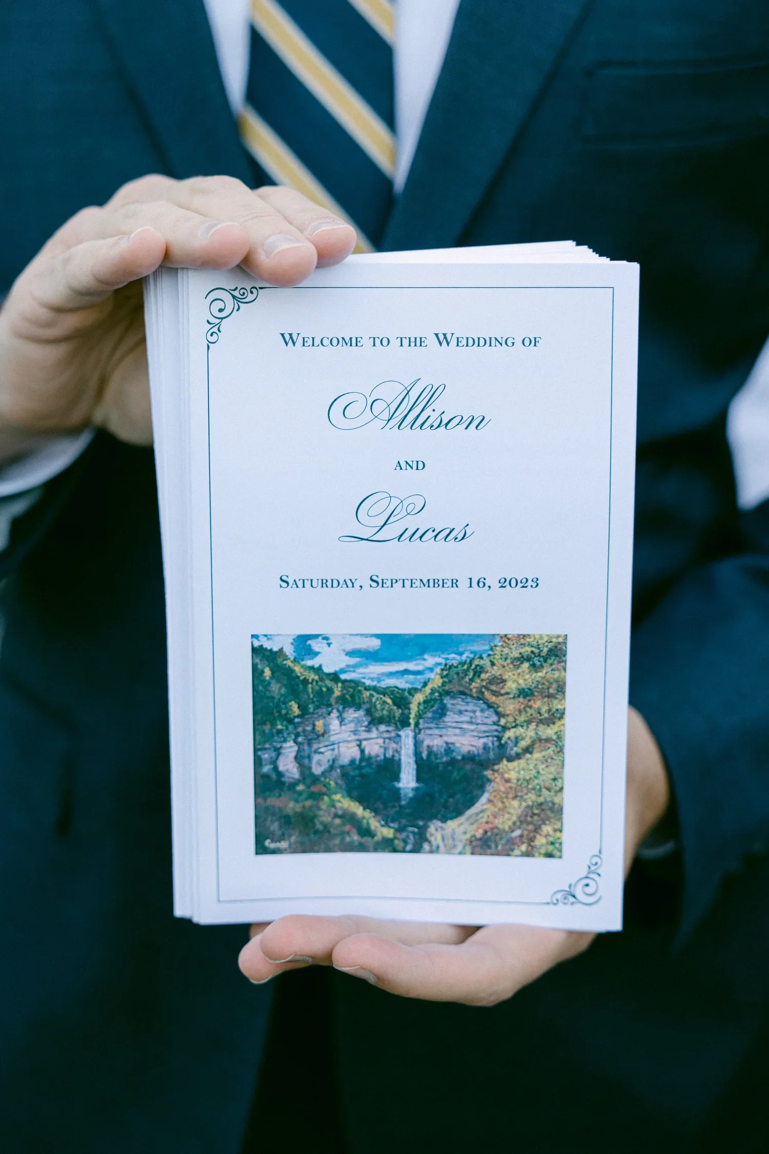 A person in a suit holding a wedding program or invitation that reads 'Welcome to the Wedding of Allison and Lucas' with the date Saturday, September 16, 2023, and includes a landscape painting of a waterfall and trees.