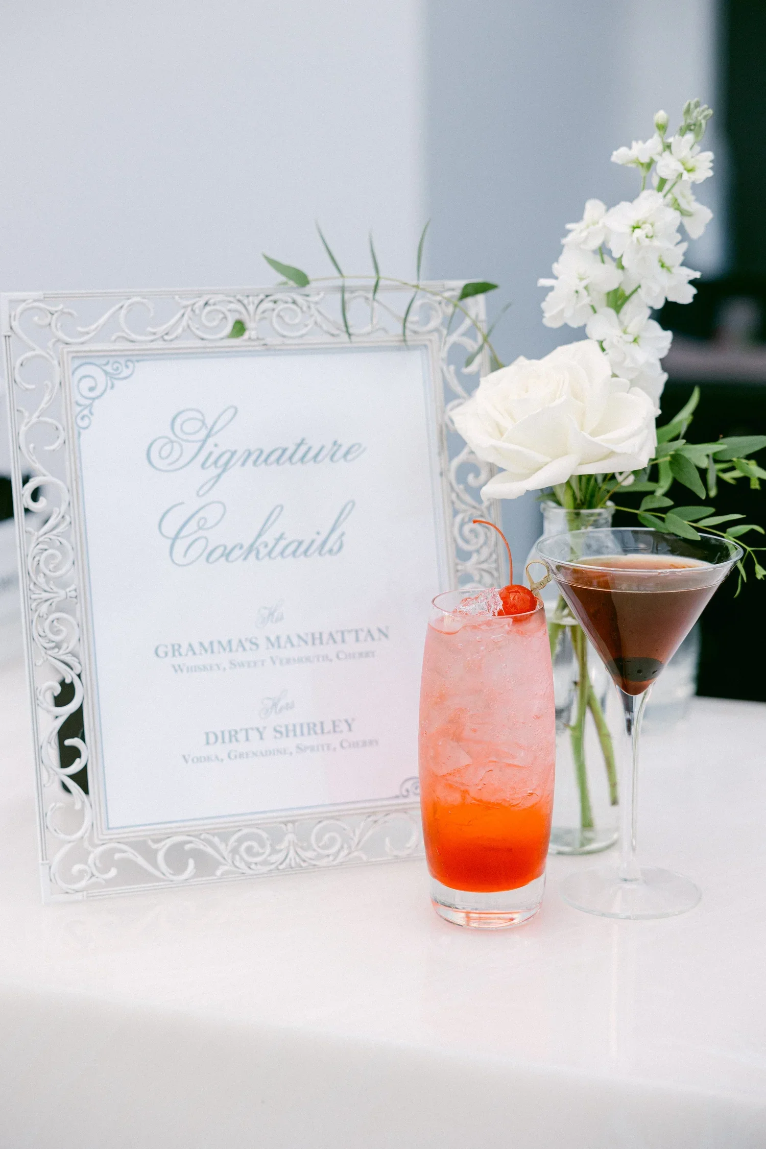 Image of a drink menu with two cocktails in front, one pink with a cherry and one dark, both in elegant glasses. A white ornate frame sign reads "Signature Cocktails" and lists two drinks: "Grammas Manhattan" and "Dirty Shirley." A white flower in a 