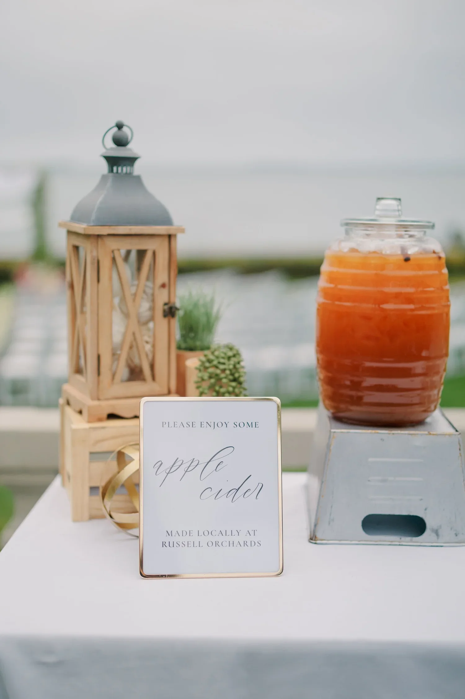 A table with a sign that reads 'Please enjoy some apple cider made locally at Russell Orchards,' a small wooden lantern, a green plant, and a large jar of apple cider, outdoors with a blurred background.