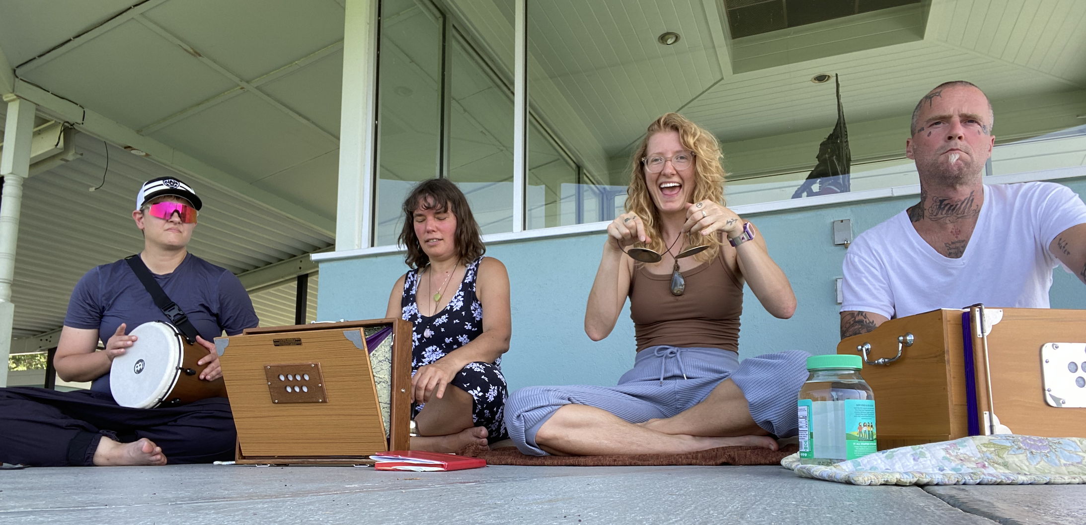 Four people sitting on a balcony, playing musical instruments and smiling, during daytime.