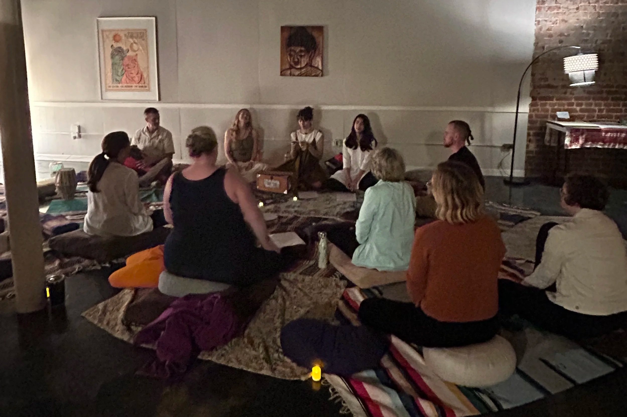 Group of people sitting on cushions and rugs in a dimly lit room, some of them engaging in a discussion or listening to a speaker at the front.