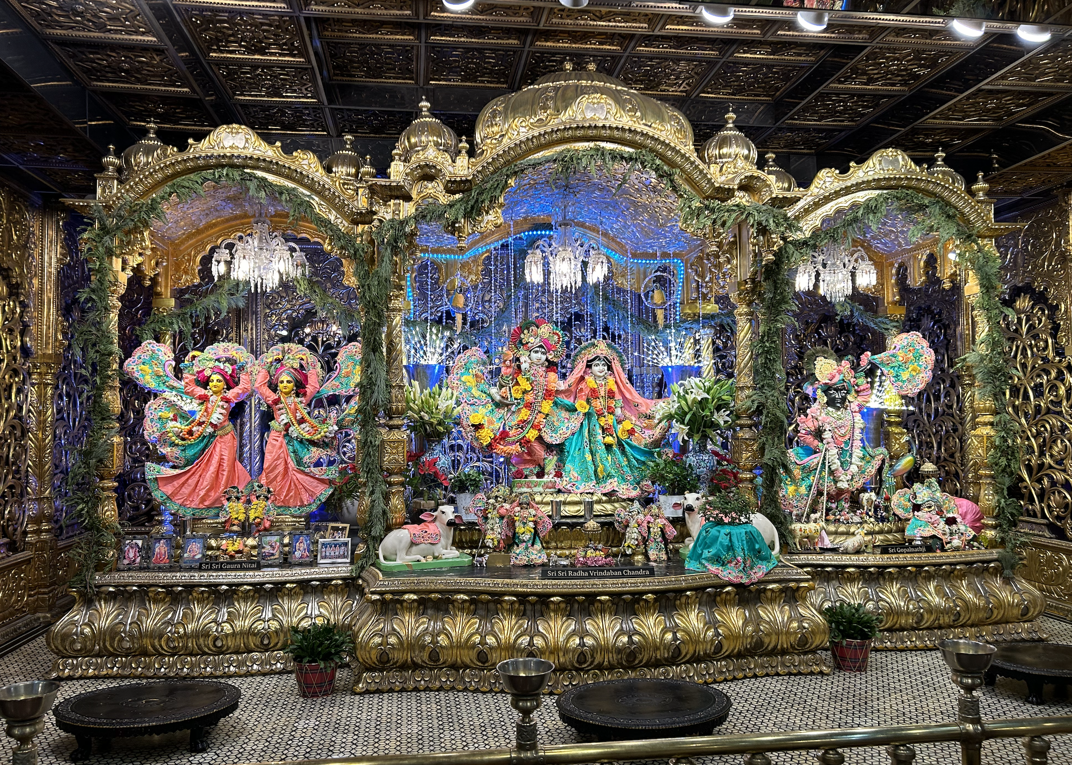Hindu altar decorated with colorful statues of deities, flowers, and greenery, with a ornate gold backdrop and chandeliers, dedicated to Sri Sri Gaura Nitai and Sri Sri Radha Vrindaban Chandra.