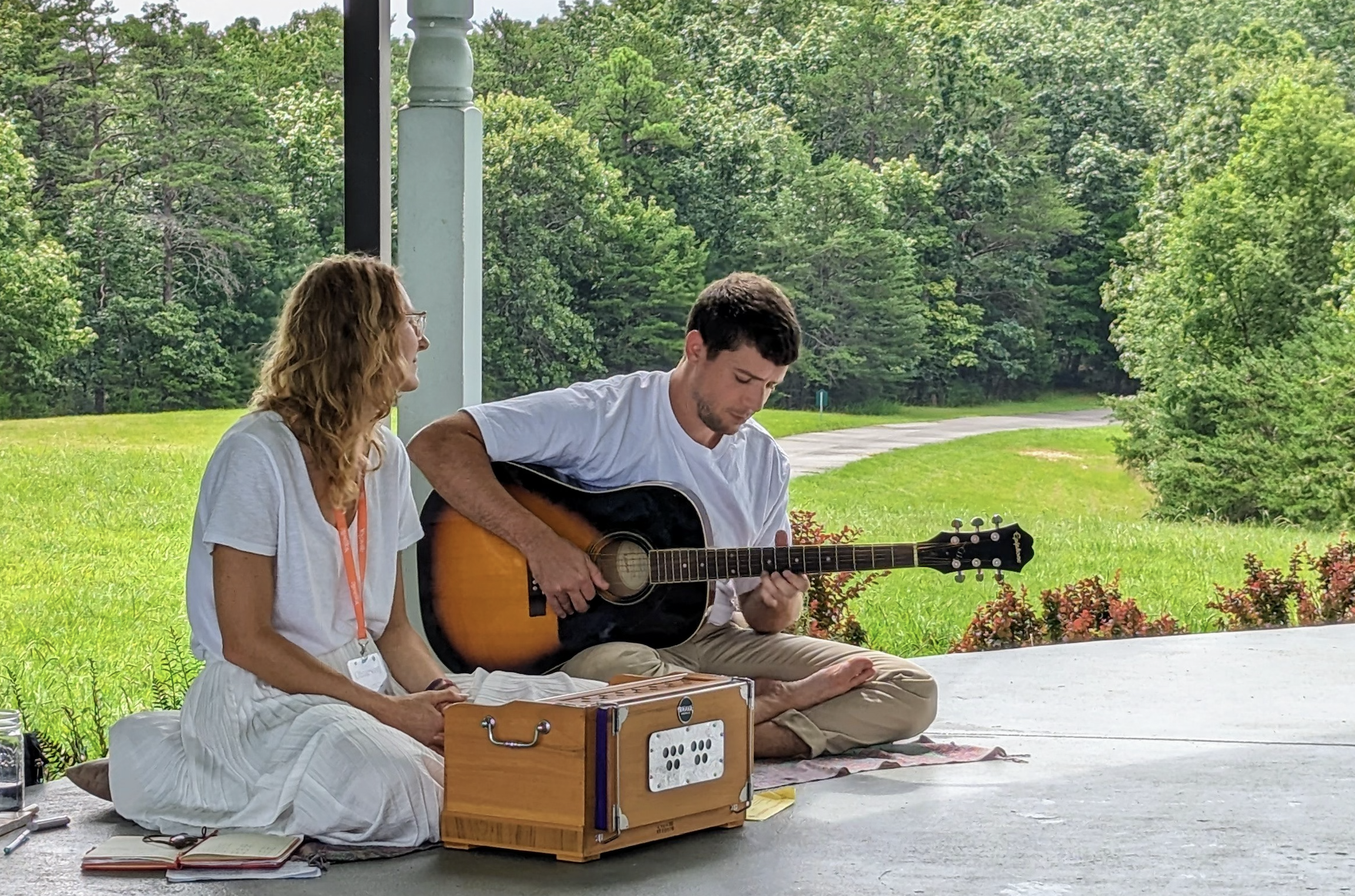 A man and a woman sitting outdoors on a grassy area, with the man playing an acoustic guitar and the woman sitting nearby. The woman is dressed in white, and the man is in light-colored clothing. They are under a covered porch, with a lush green fore