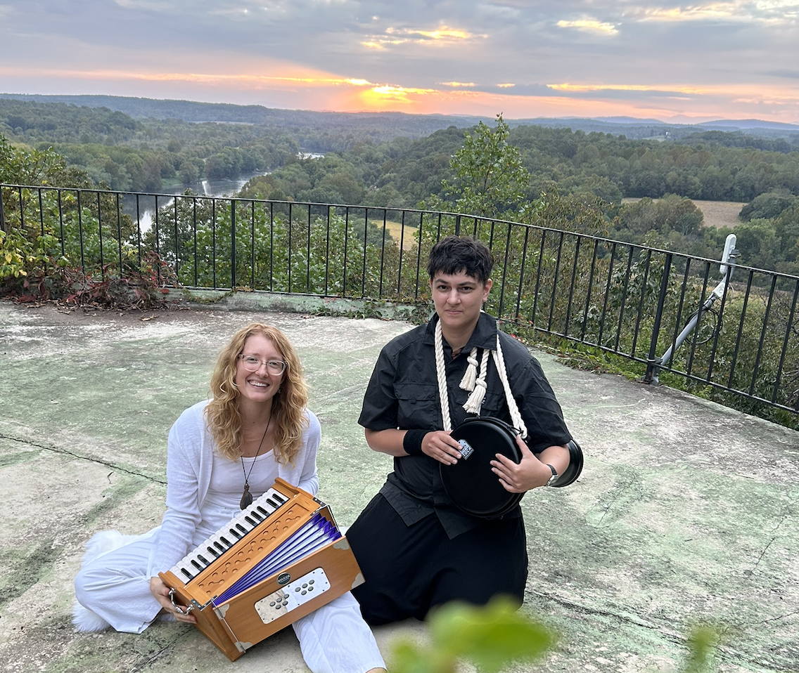Two women sitting outdoors on a concrete surface during sunset, with scenic trees and a river in the background. One woman is smiling, holding a melodica, and the other has a serious expression, holding a drum and standing near a railing.