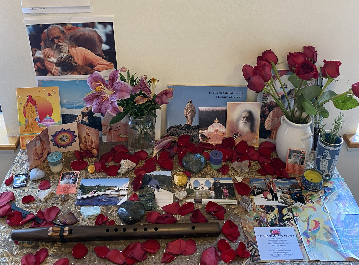 A spiritual altar featuring flowers, photographs, candles, rocks, and various religious and spiritual items. Red rose petals are scattered across the surface, with pink lilies and separate rose bouquets in vases. Pictures of spiritual leaders and sce