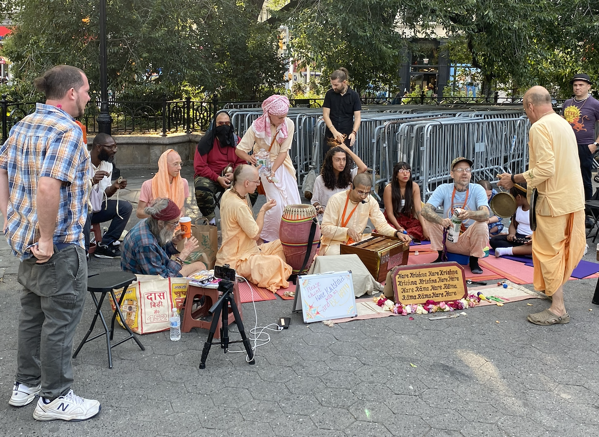 Hare Krishna devotees gathered outdoors, sitting on mats and playing musical instruments, with some people standing around. There are signs and flowers in front, and metal barricades in the background.