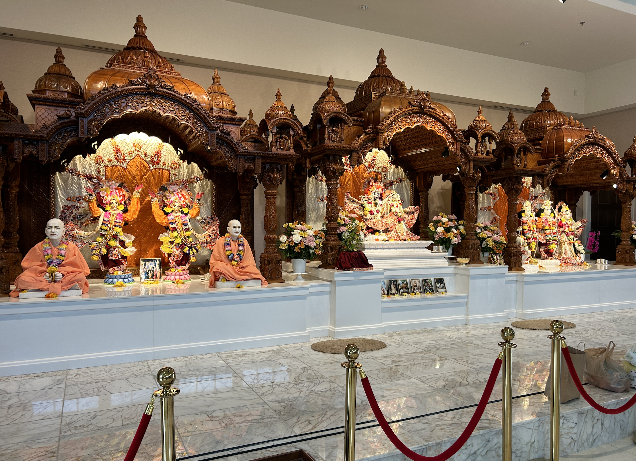 Hindu temple altar with colorful sculptures of deities, framed by intricate wooden architecture, and surrounded by flowers and photographs.