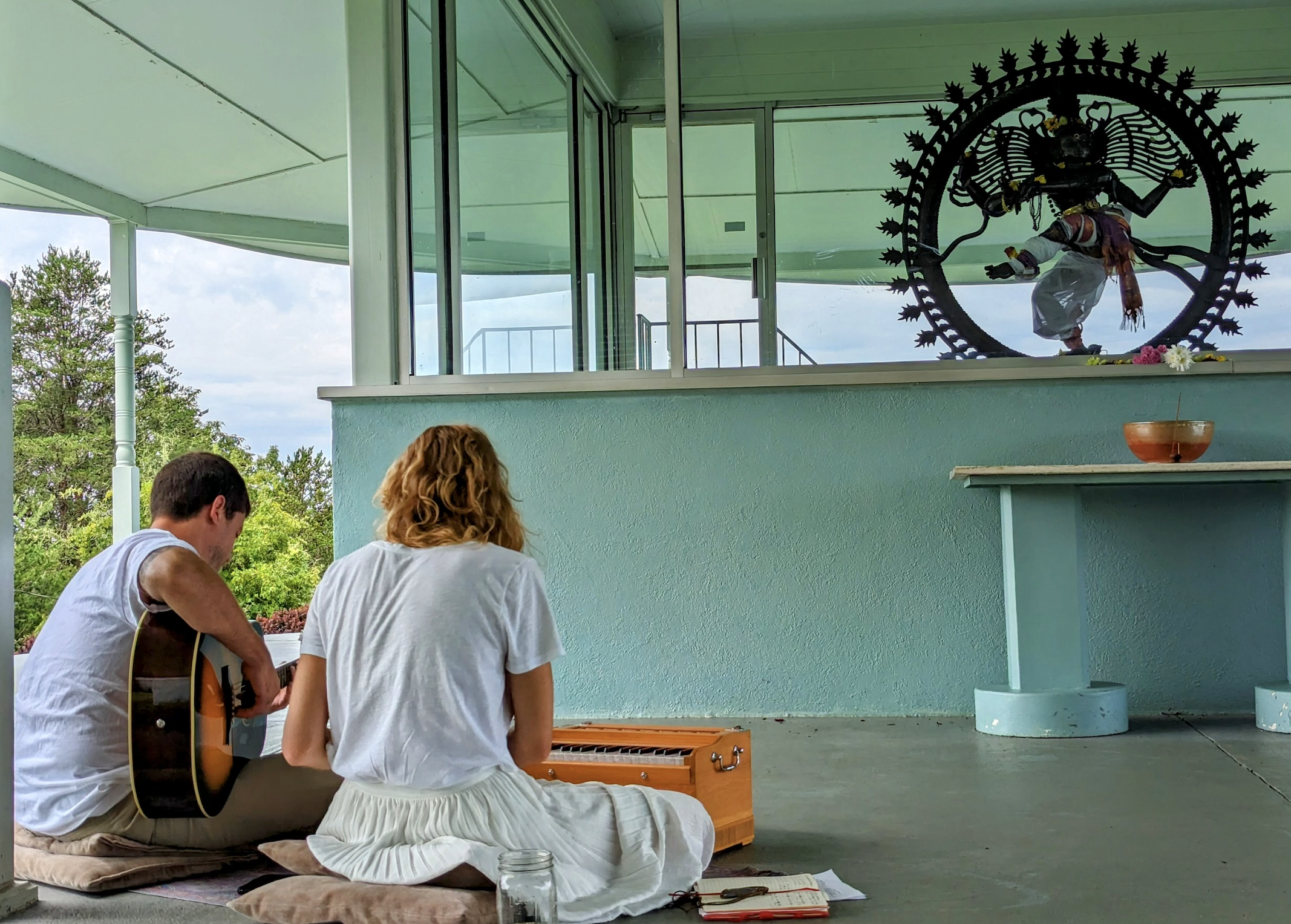 A musician playing guitar and singing to a woman who is sitting and listening in an outdoor covered area. There is a statue of a multi-armed figure with a circular frame behind it on the wall.