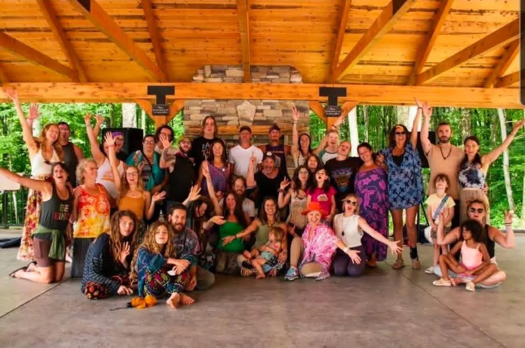 Group of people in colorful clothing posing under a wooden pavilion with a stone fireplace, surrounded by green trees.