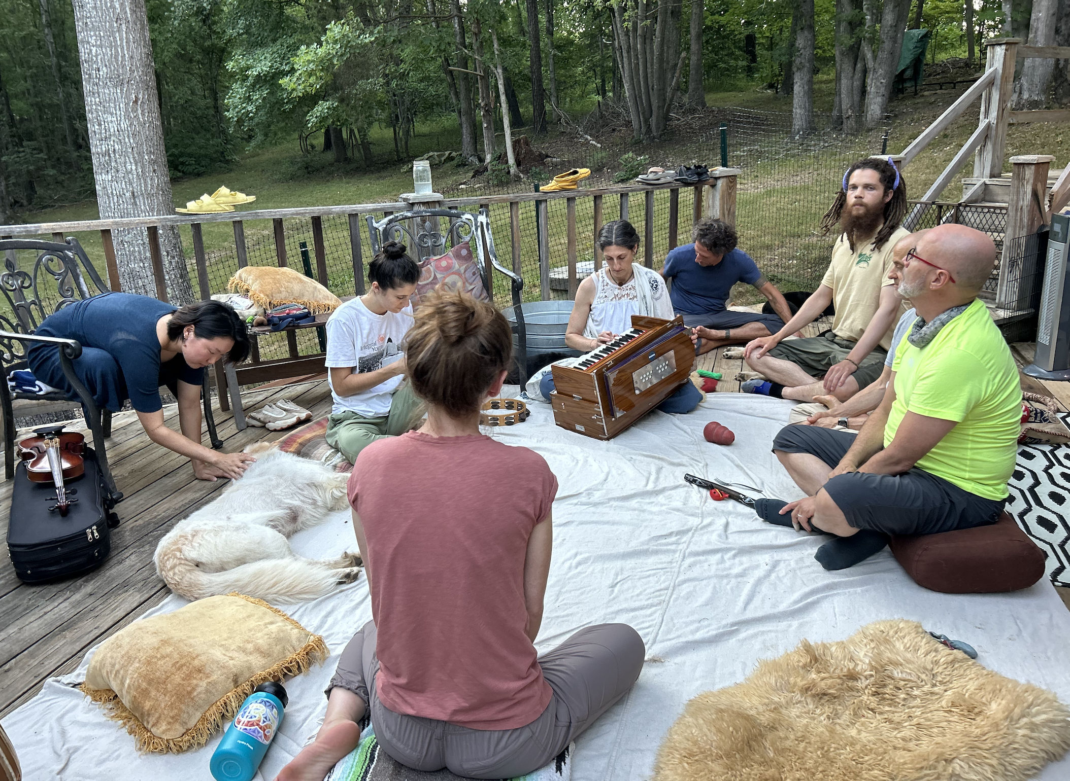 A group of people sitting and kneeling on a wooden deck outdoors, with a forested background. They are gathered around a small portable piano, some are playing or looking at it. There are pillows, shoes, and a dog lying down on a blanket in the foreg