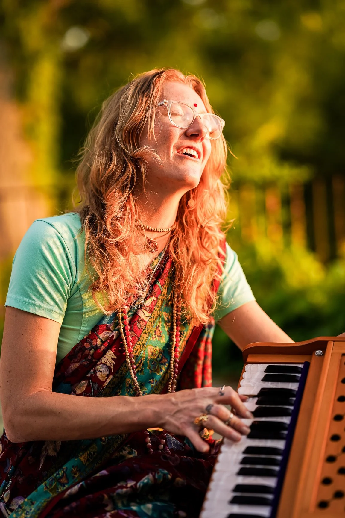 A woman with long, wavy red hair and glasses playing a small keyboard outdoors, smiling with eyes closed, wearing a green shirt, patterned scarf, and beads, with sunlight and trees in the background.