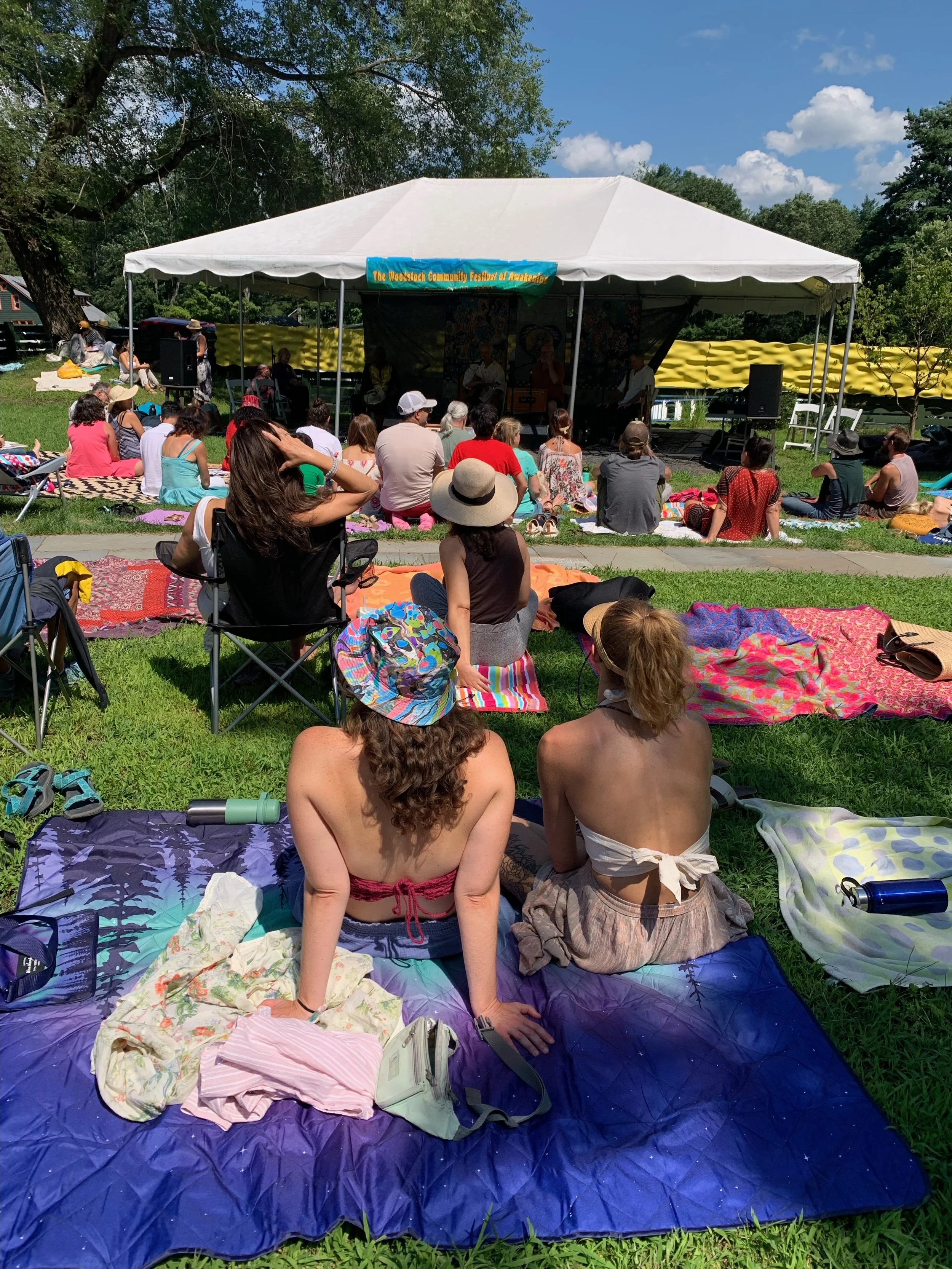 An outdoor summer festival with people sitting on blankets and chairs near a white canopy tent, with a stage and performers, surrounded by green trees and blue sky.