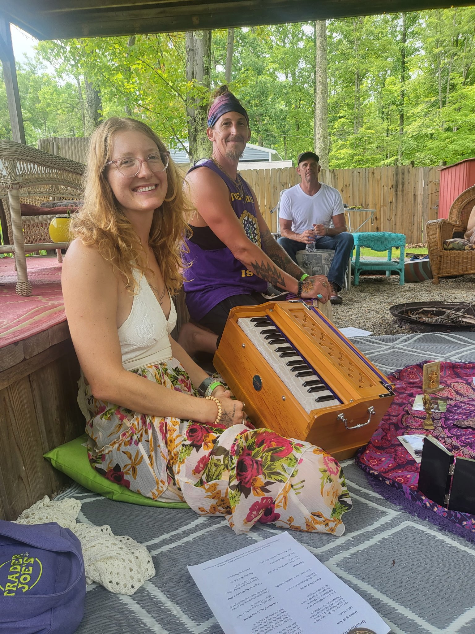 Three people sitting outdoors on a covered patio area surrounded by trees and greenery. One woman with glasses and long, wavy blonde hair is smiling at the camera, wearing a white top and floral skirt, sitting on a green cushion with a yellow and ora