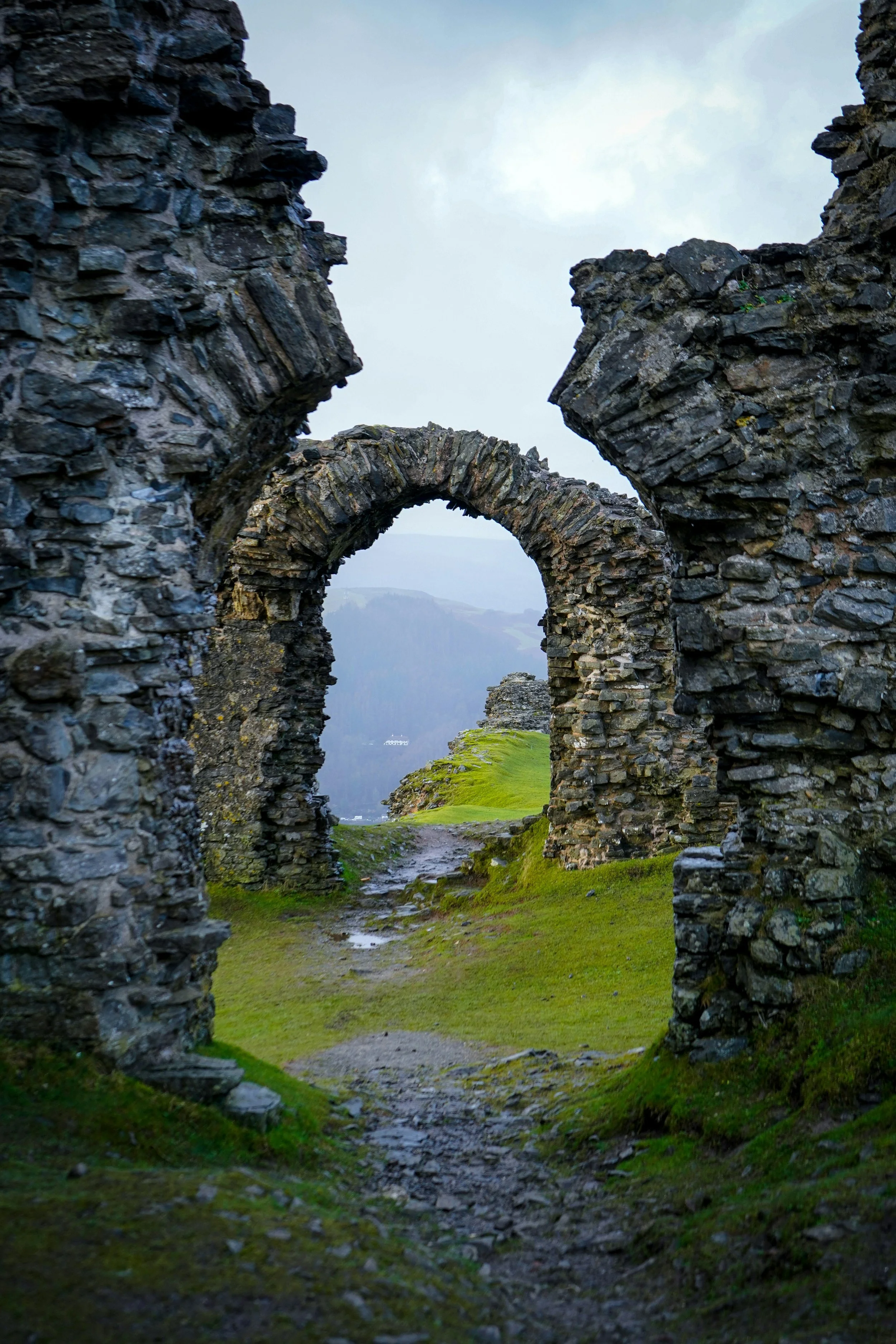 Ancient stone ruins with arches on a grassy hill, overlooking a misty landscape with mountains in the background.