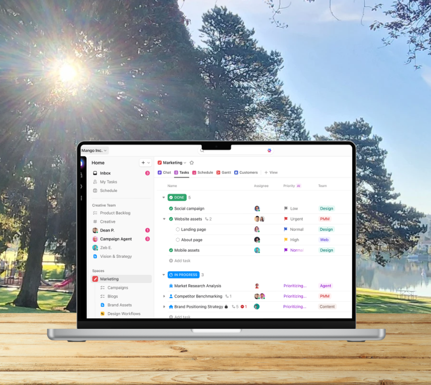 A laptop displaying a marketing project management interface outdoors on a wooden surface with trees and a lake in the background and the sun shining through the trees.